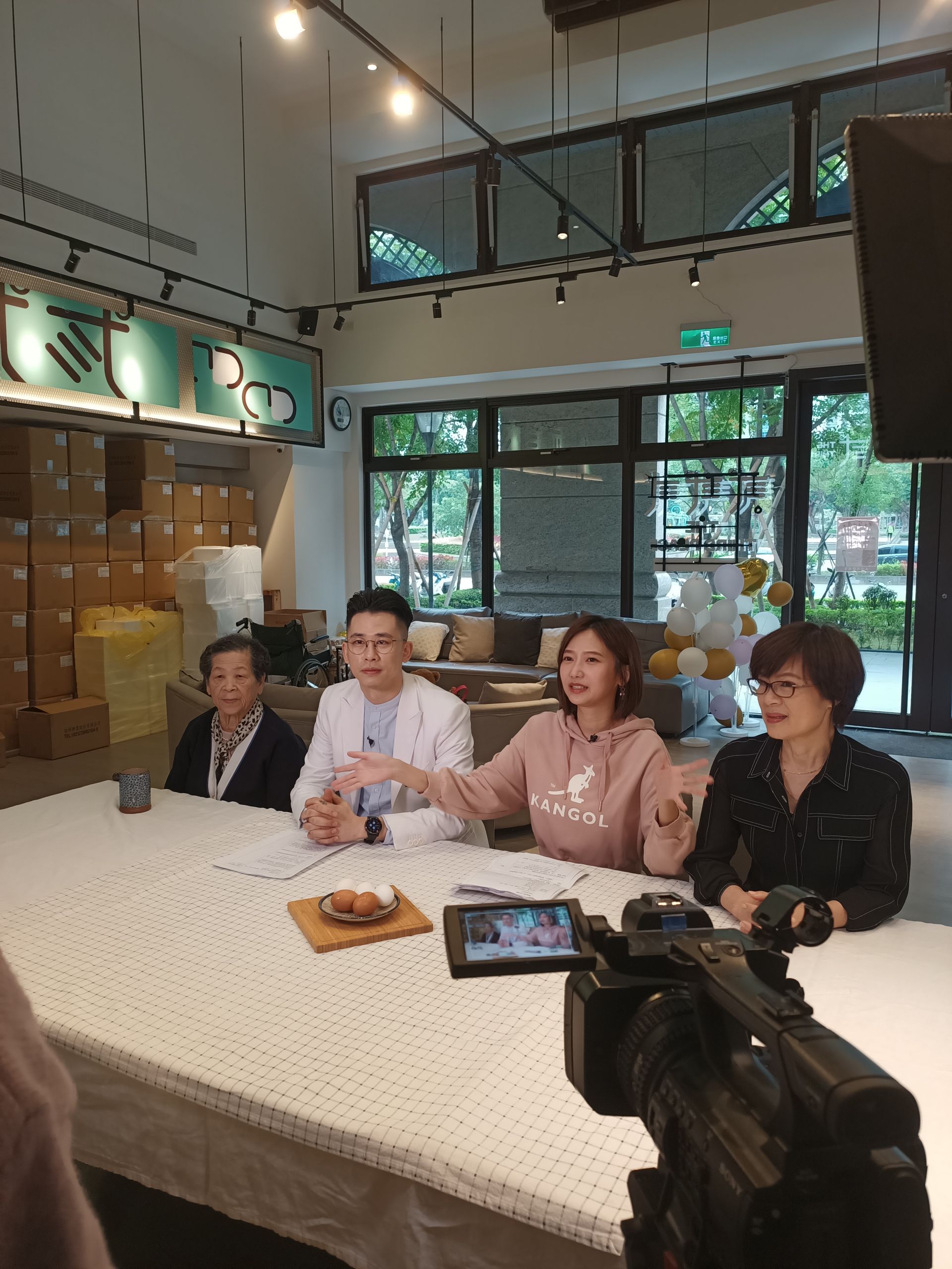 Four people seated at a table, discussing, with a video camera. Inside a brightly lit room, with boxes in the background.
