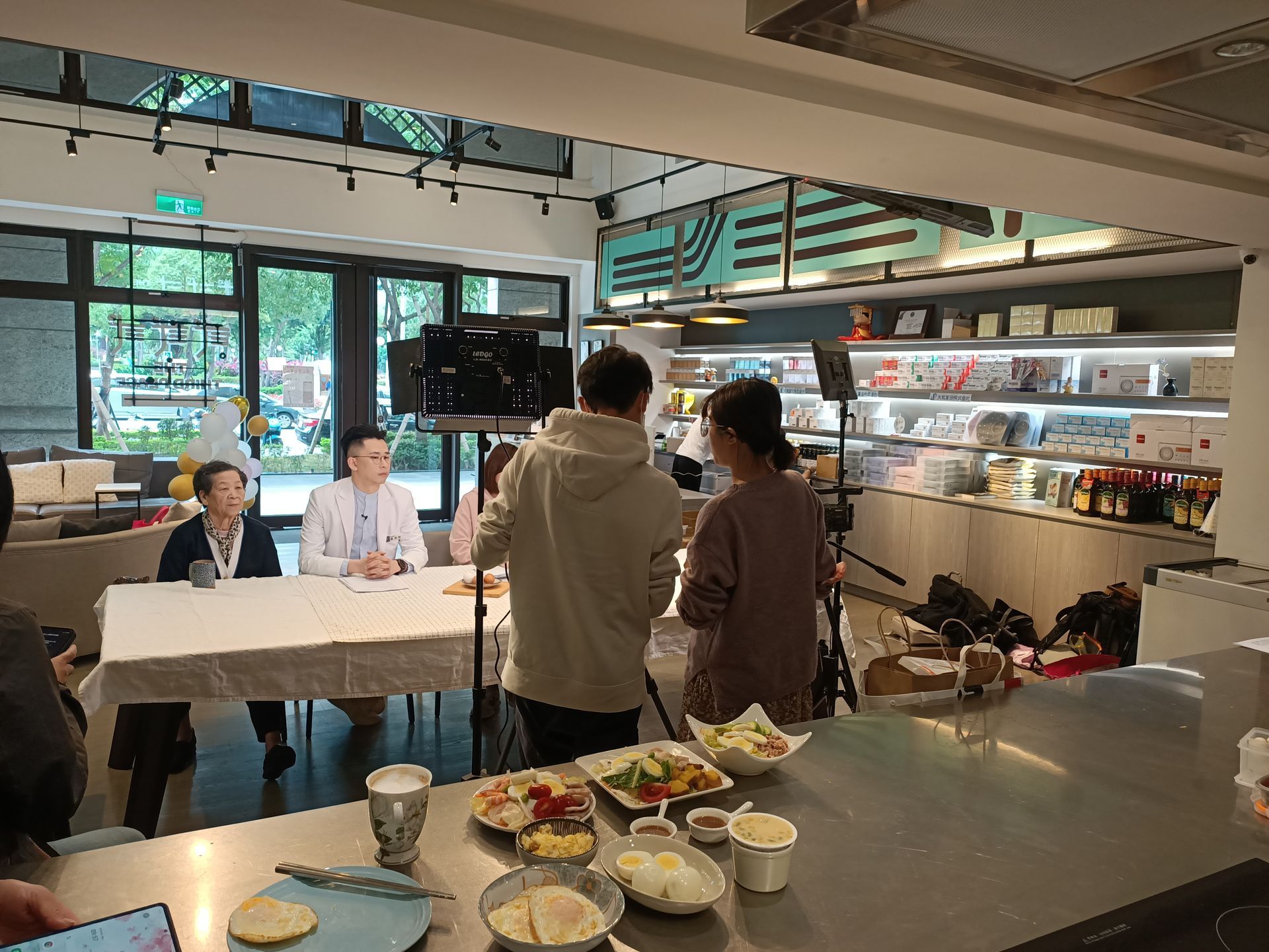 Two people at table with microphones, being filmed in a store with food on counter.
