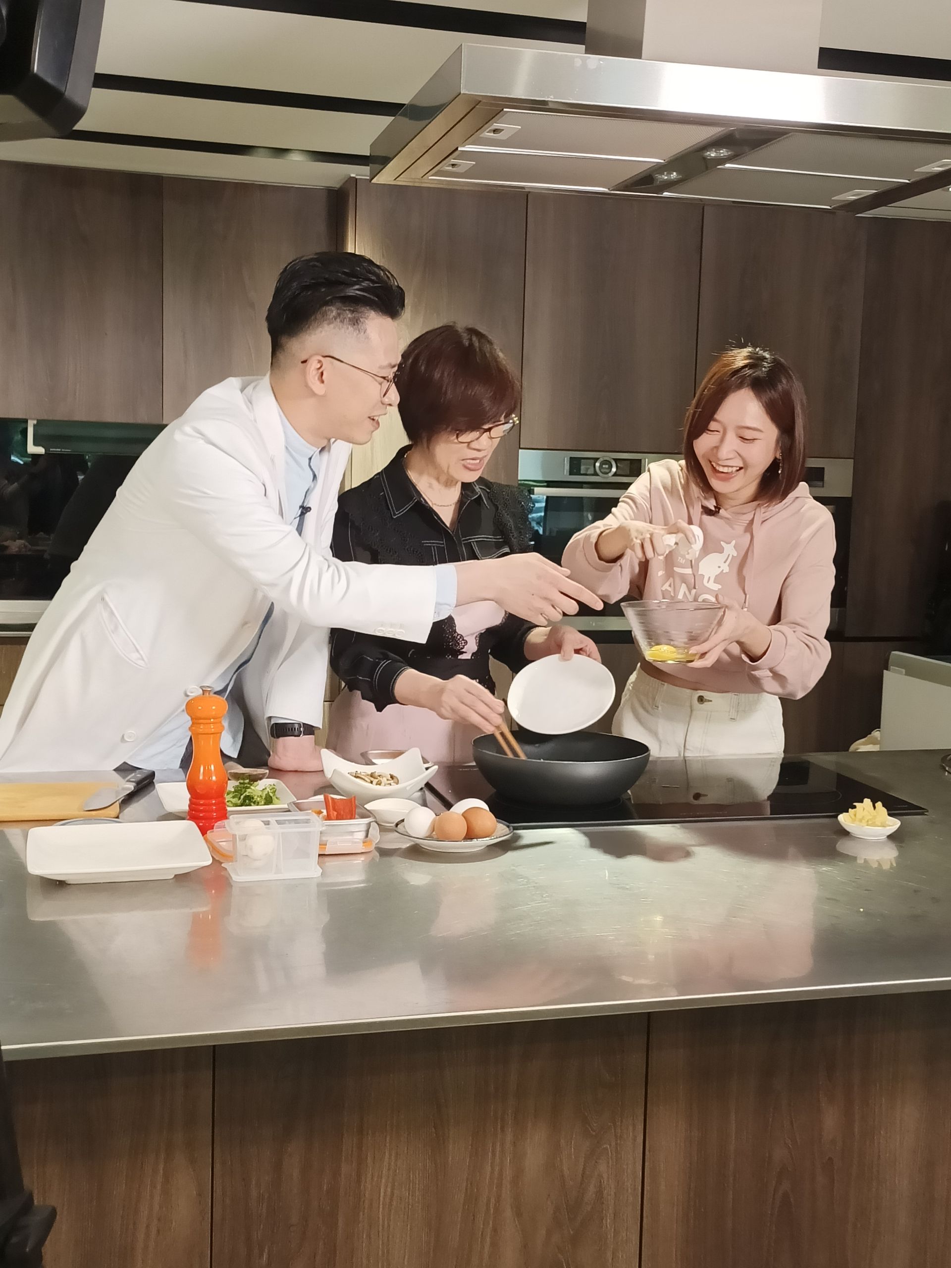 Three people cooking together in a modern kitchen, smiling. Woman pours ingredients into a black wok.