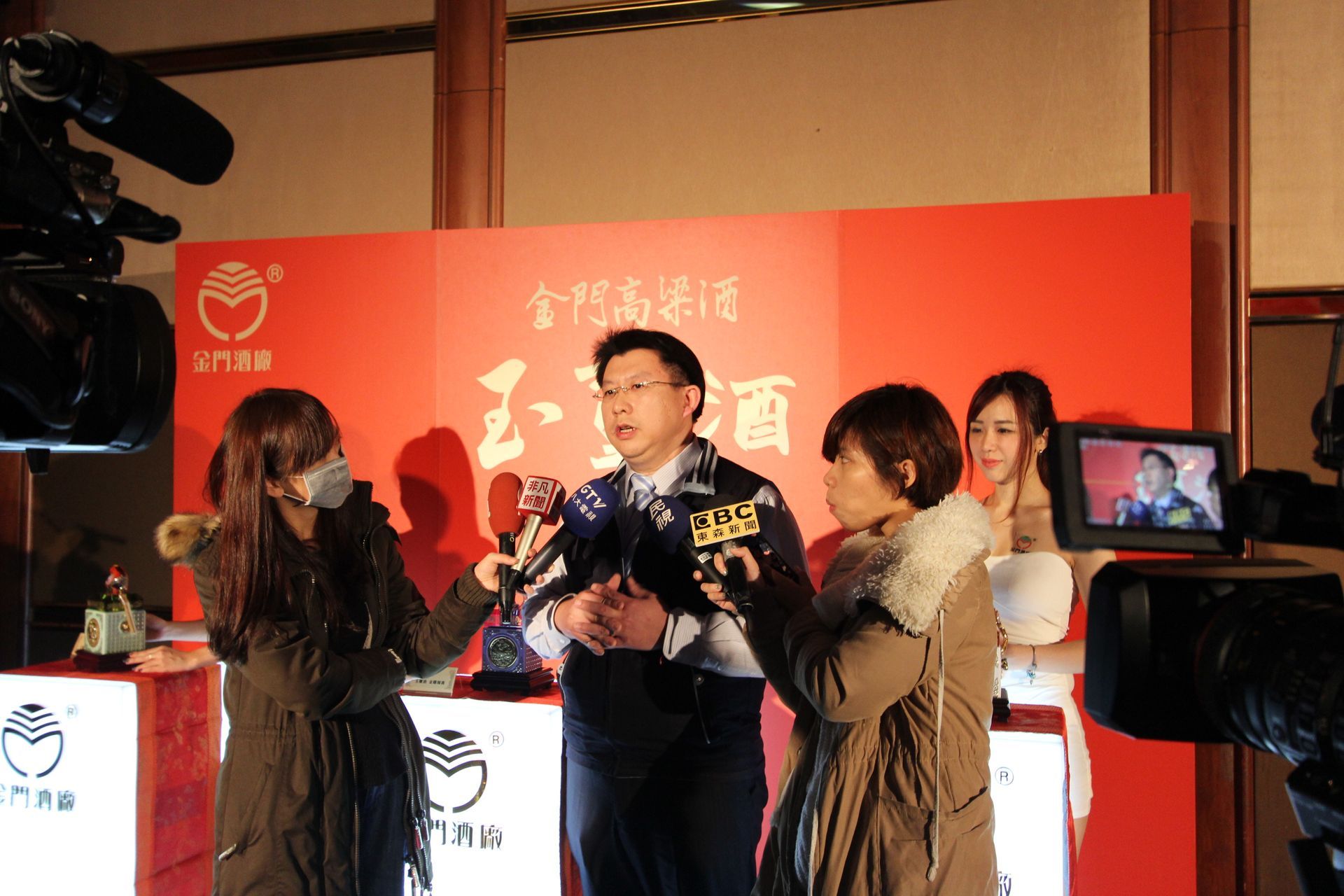 Man speaking to reporters with microphones at a red backdrop event. Several cameras and monitors are present.