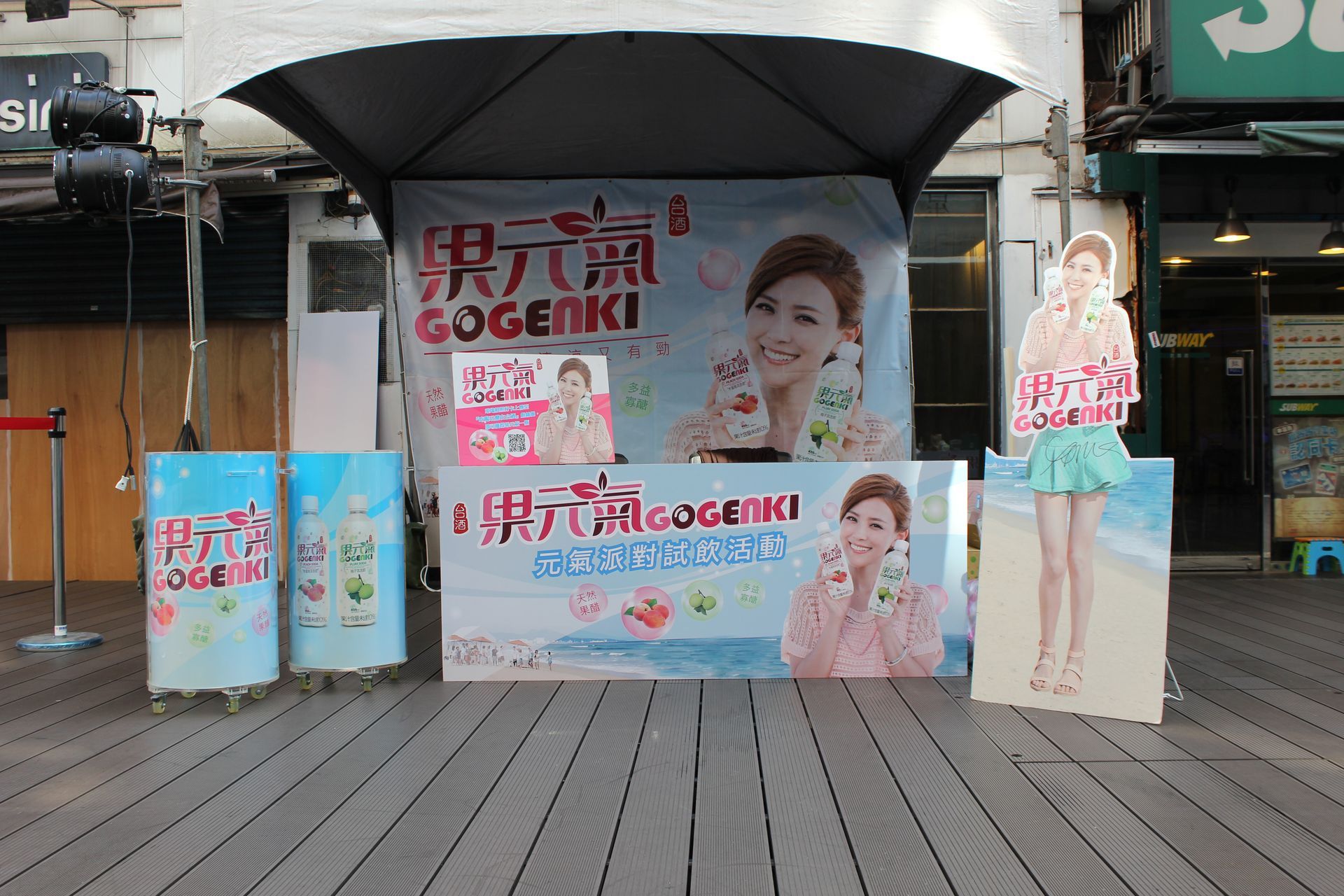 Promotional stand with a woman advertising a drink, displayed in front of a building with a wooden deck.