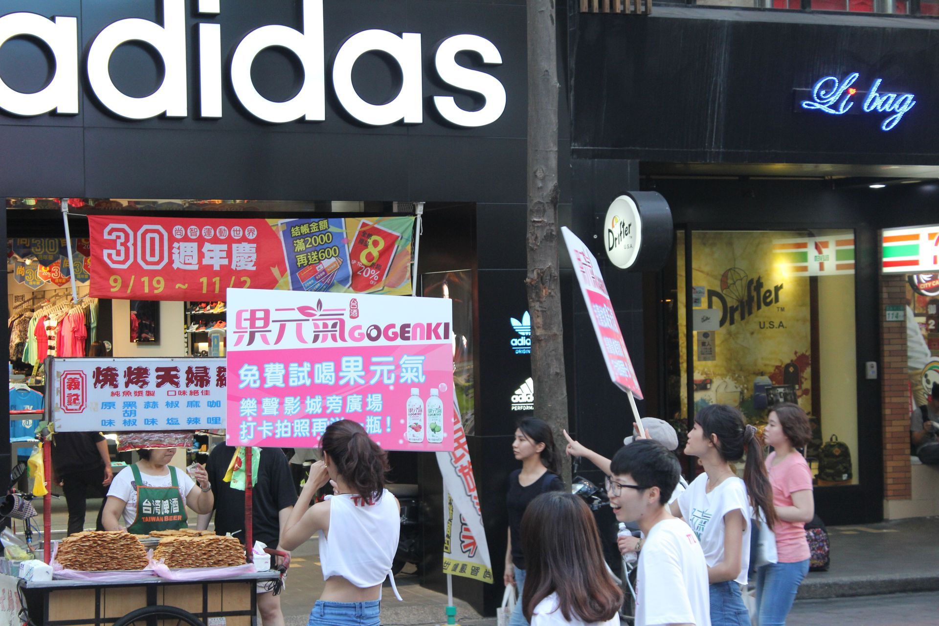Street scene with an Adidas store, food stall, and people. A sign advertises 