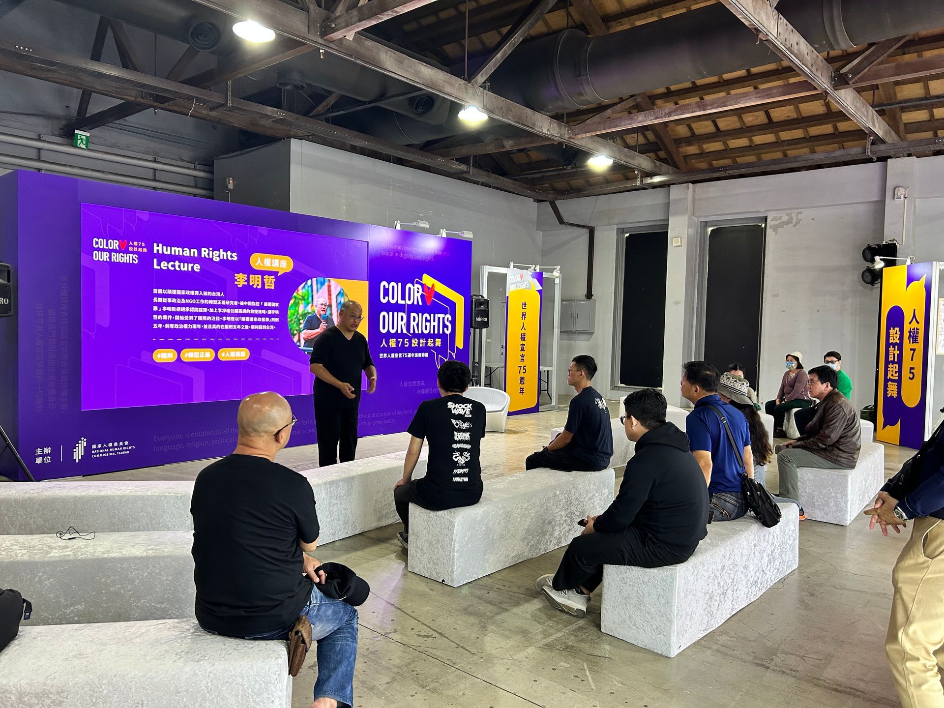 Man giving presentation on stage, audience seated on blocks. Purple backdrop, modern interior.