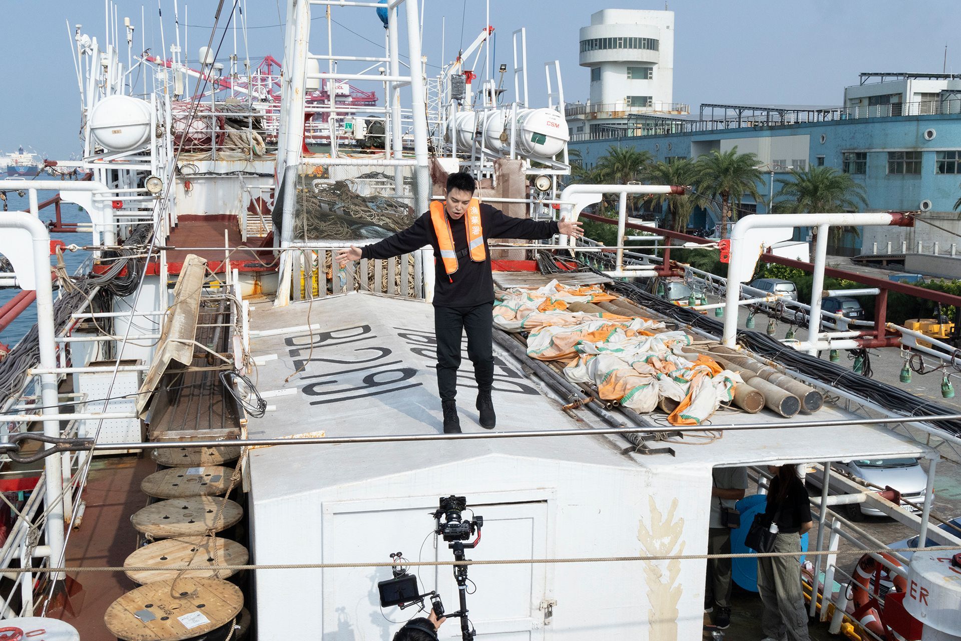 Man balancing on a wire on a ship, arms outstretched. He wears black and an orange life vest.