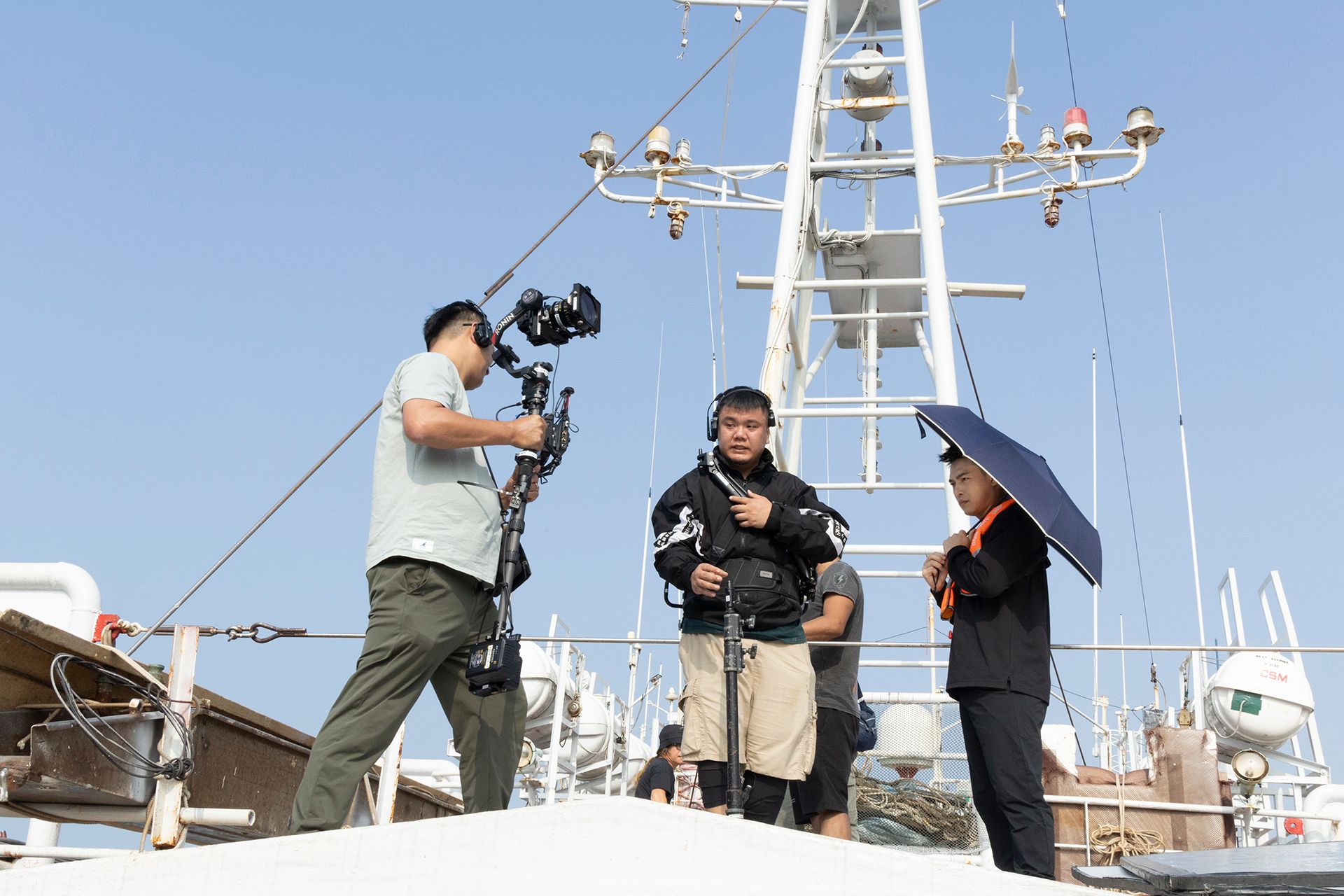 Filming a scene on a boat deck. Crew members with camera equipment, a parasol, and the ship's mast. Sunny day.