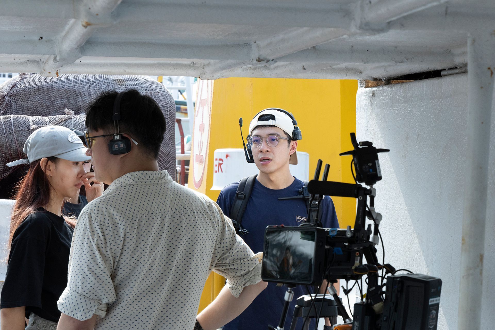Film crew on a boat filming. Two wear headsets, camera on a tripod, yellow wall in the background.