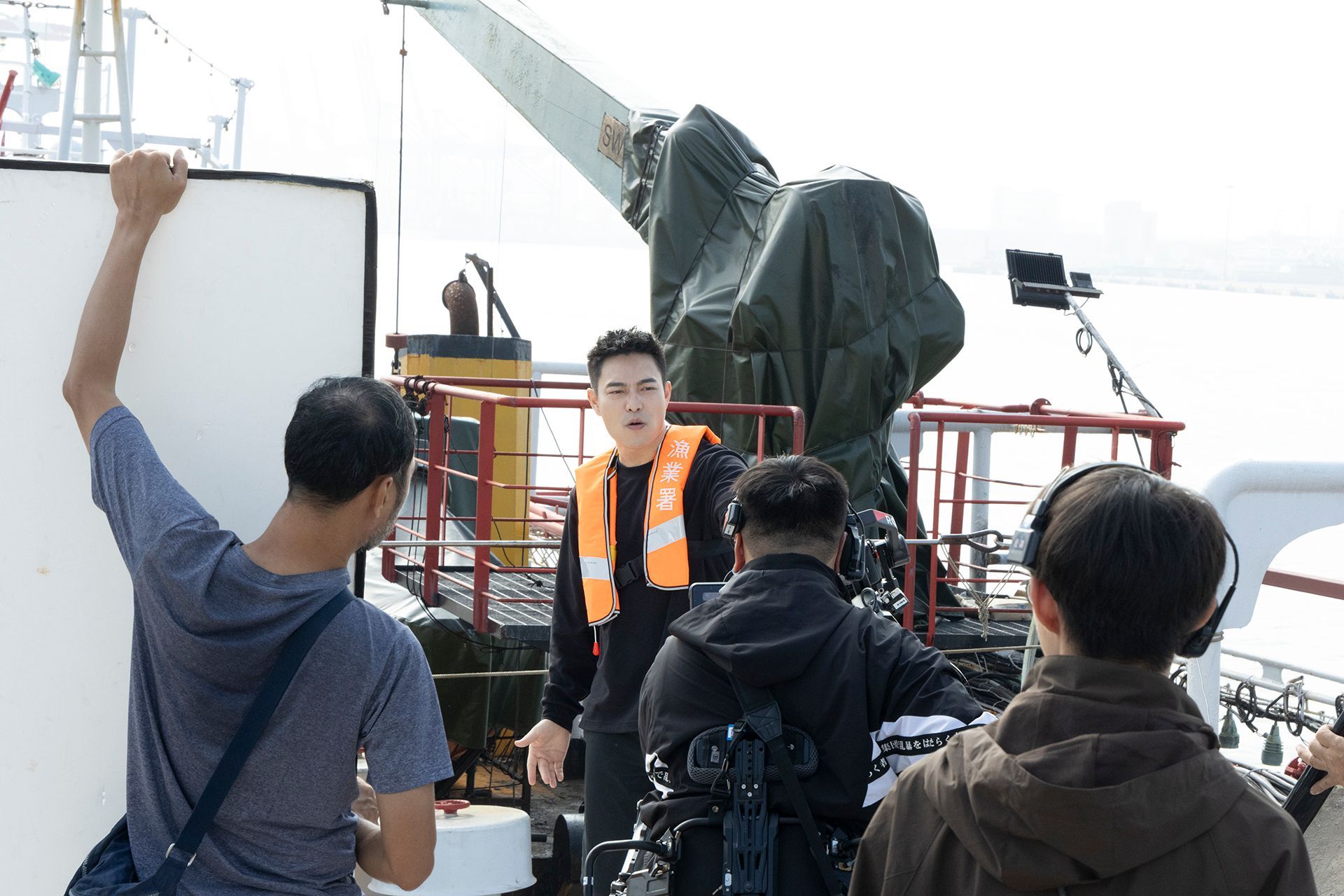 Crew filming a man on a boat; man wearing life vest gestures. Equipment and crew surround him on deck.