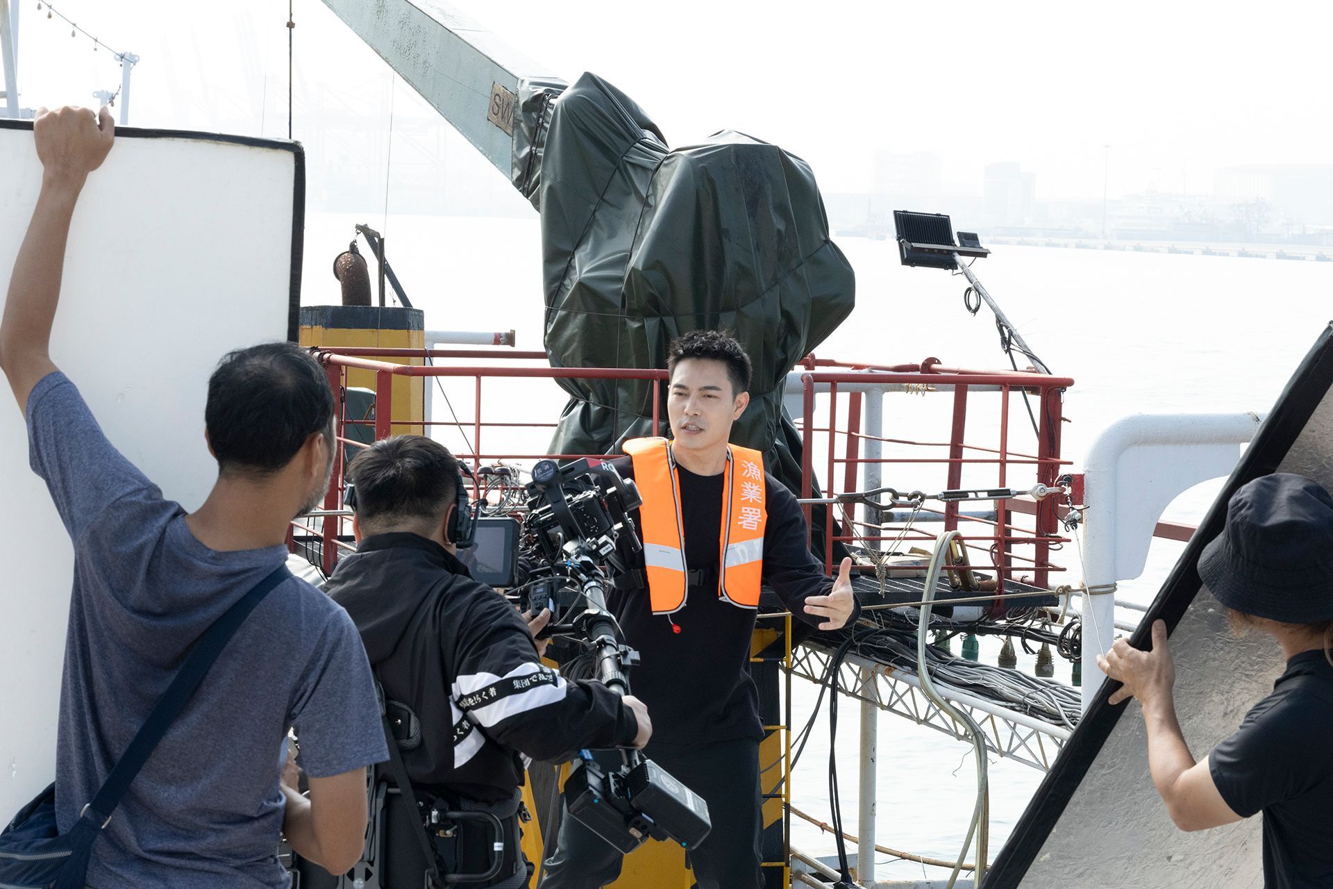 Man in orange life vest speaks on a boat deck, surrounded by film crew and equipment.