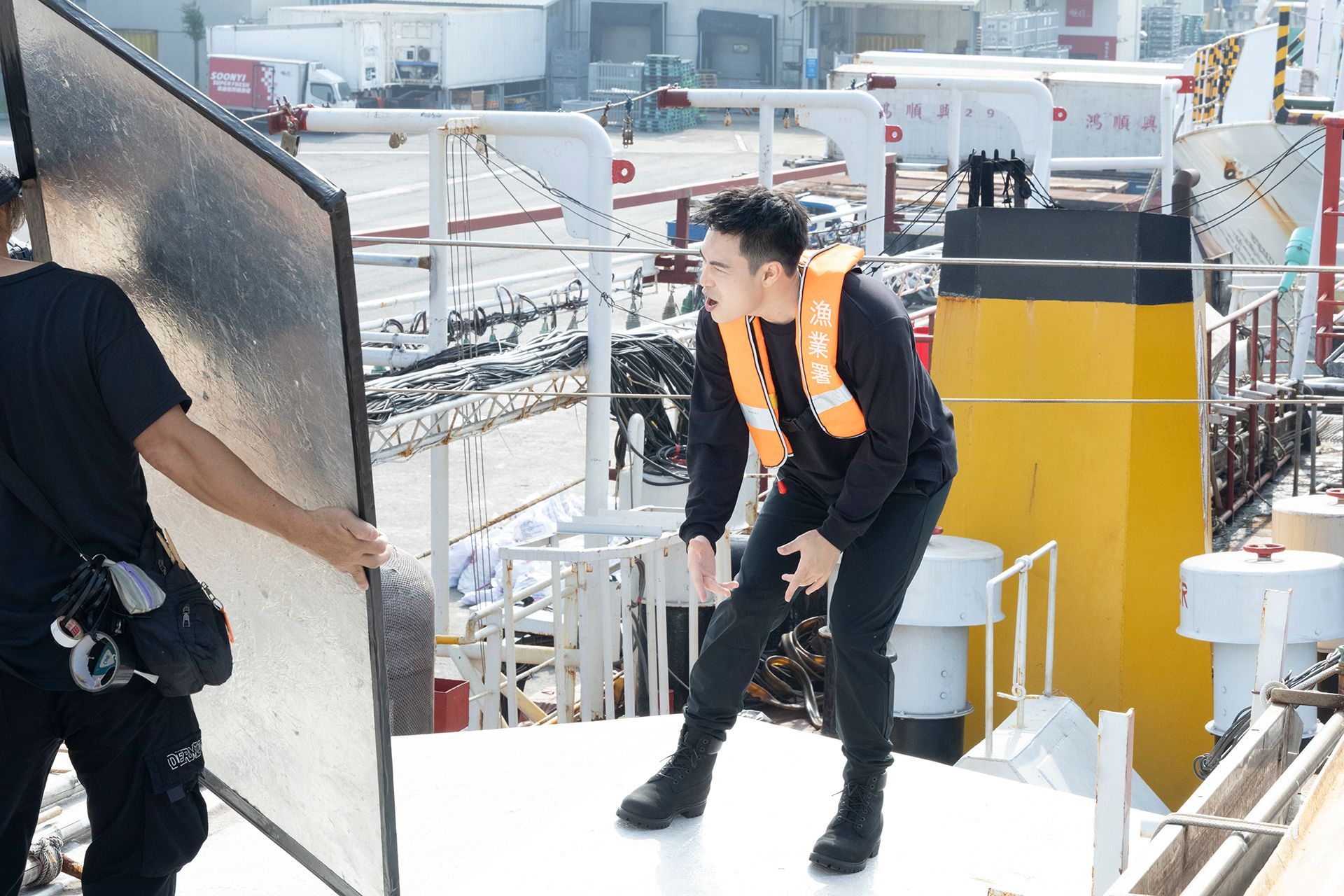 Man on ship deck with orange life vest, looking off to the side, and a crew member holding a reflector.