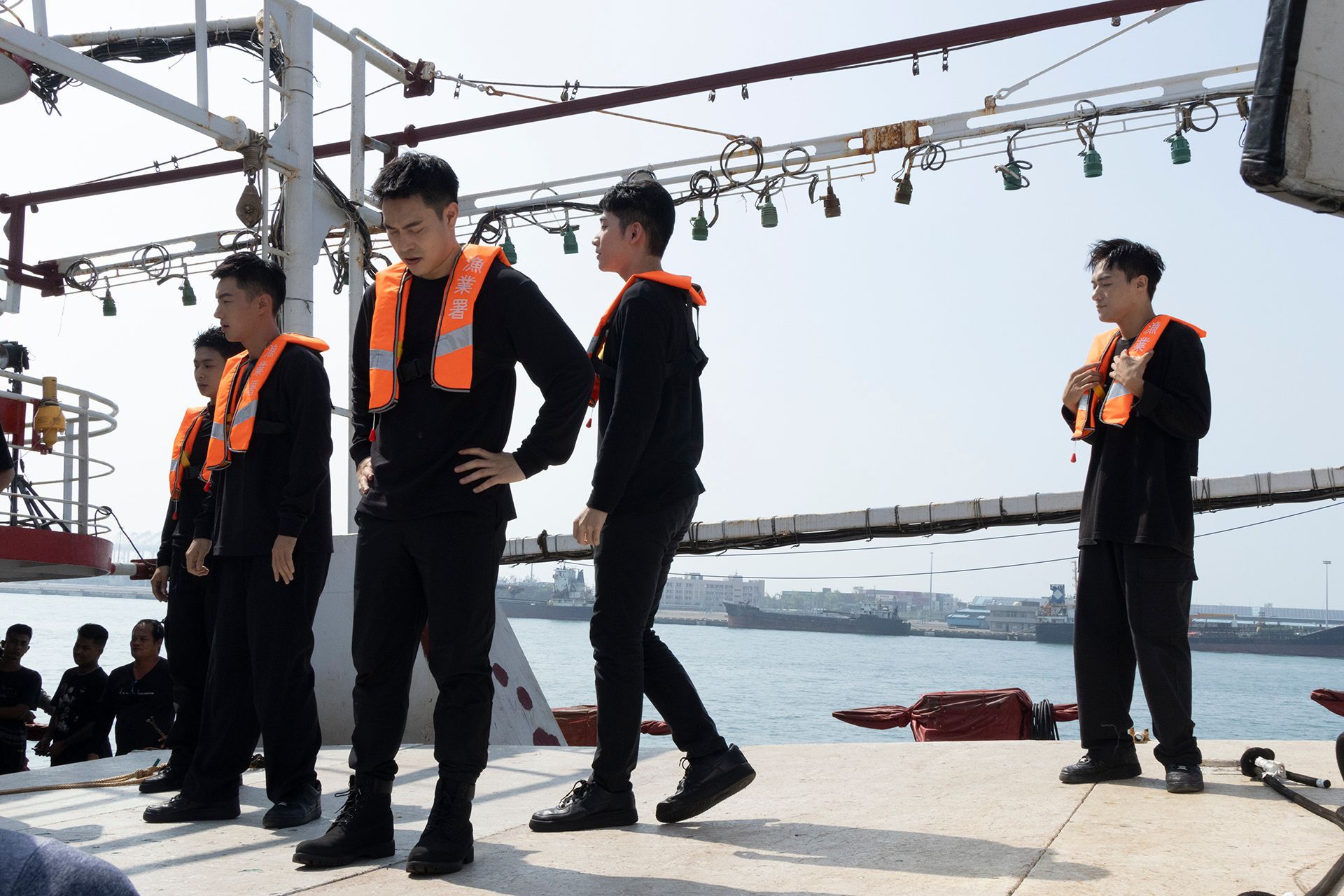 Men in black uniforms and orange life vests on a boat deck, standing near the water.