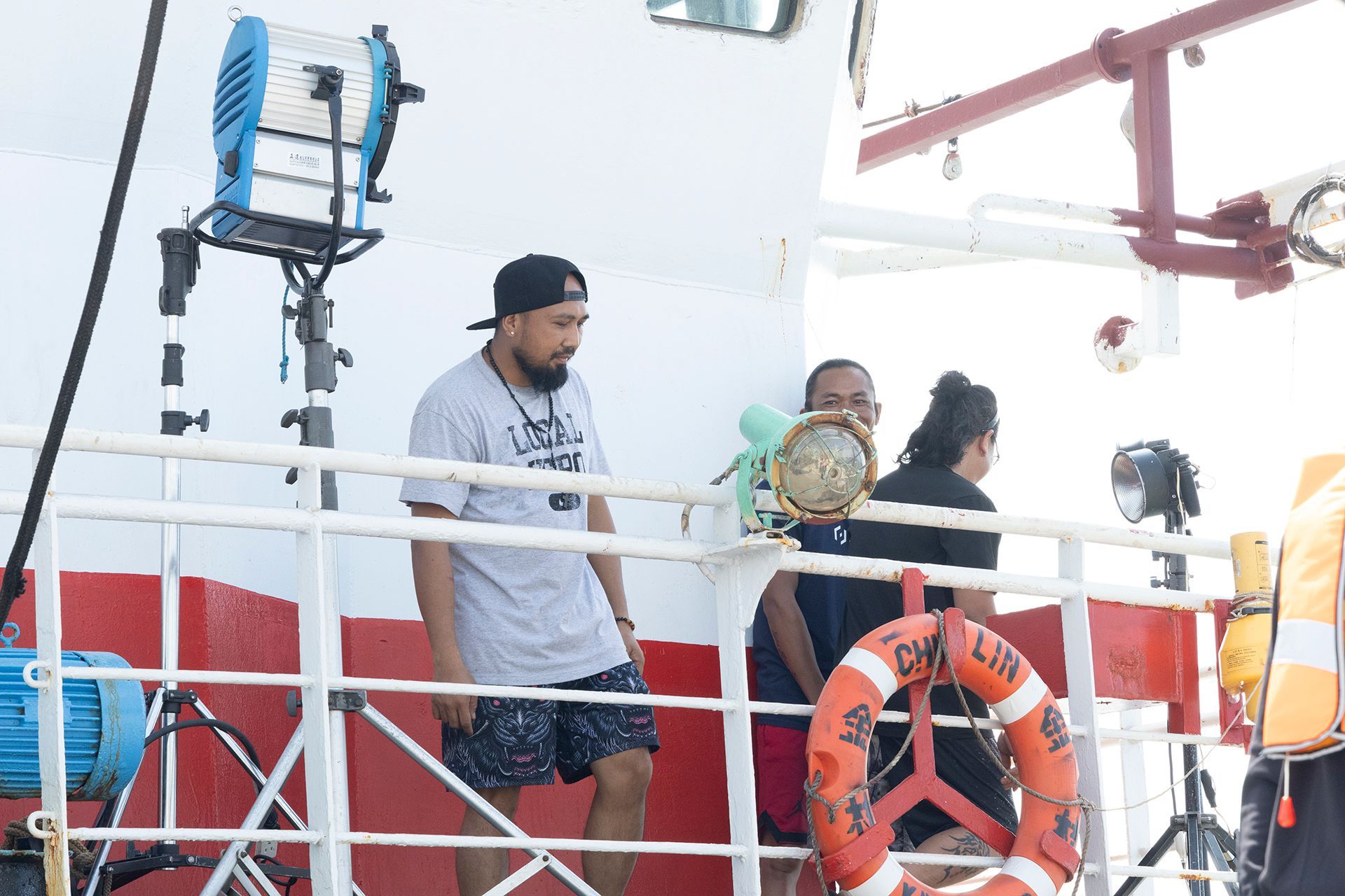 Crew on a ship deck setting up for filming. Blue light, life preservers and red details visible.