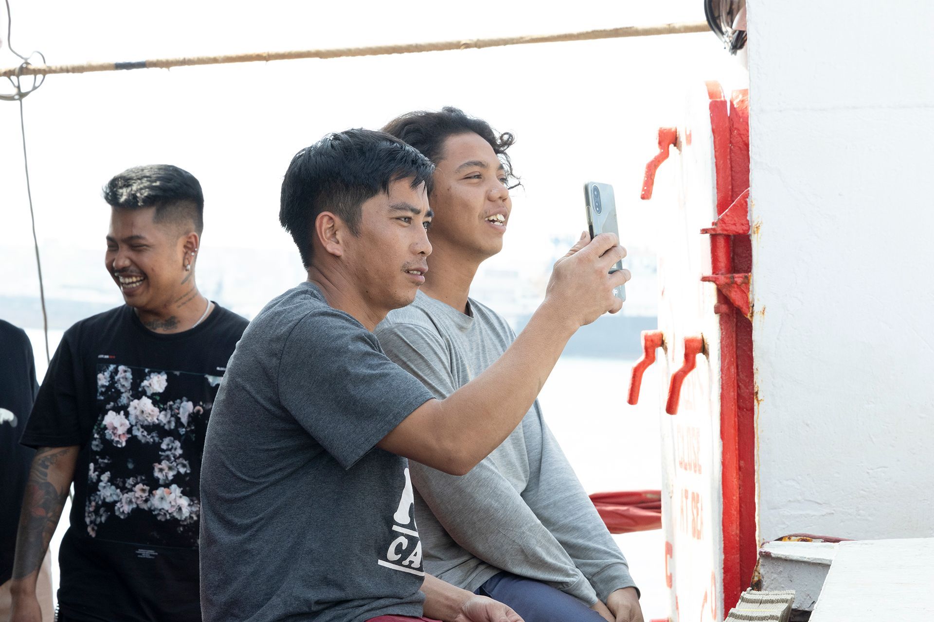 Two men taking a selfie on a boat; another man laughs in the background. Bright sunlight.
