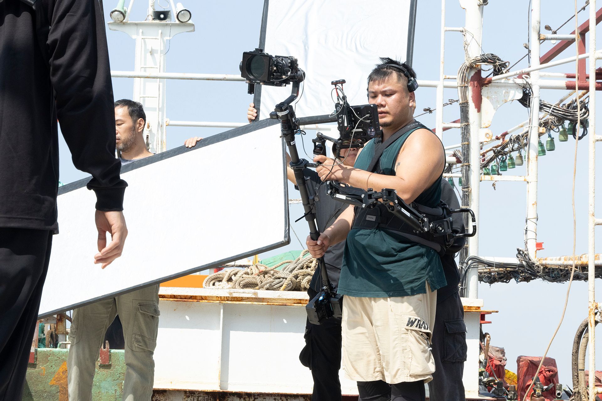 Film crew on a ship: a person with a camera, another holding a white reflector, and a third holding a black object.