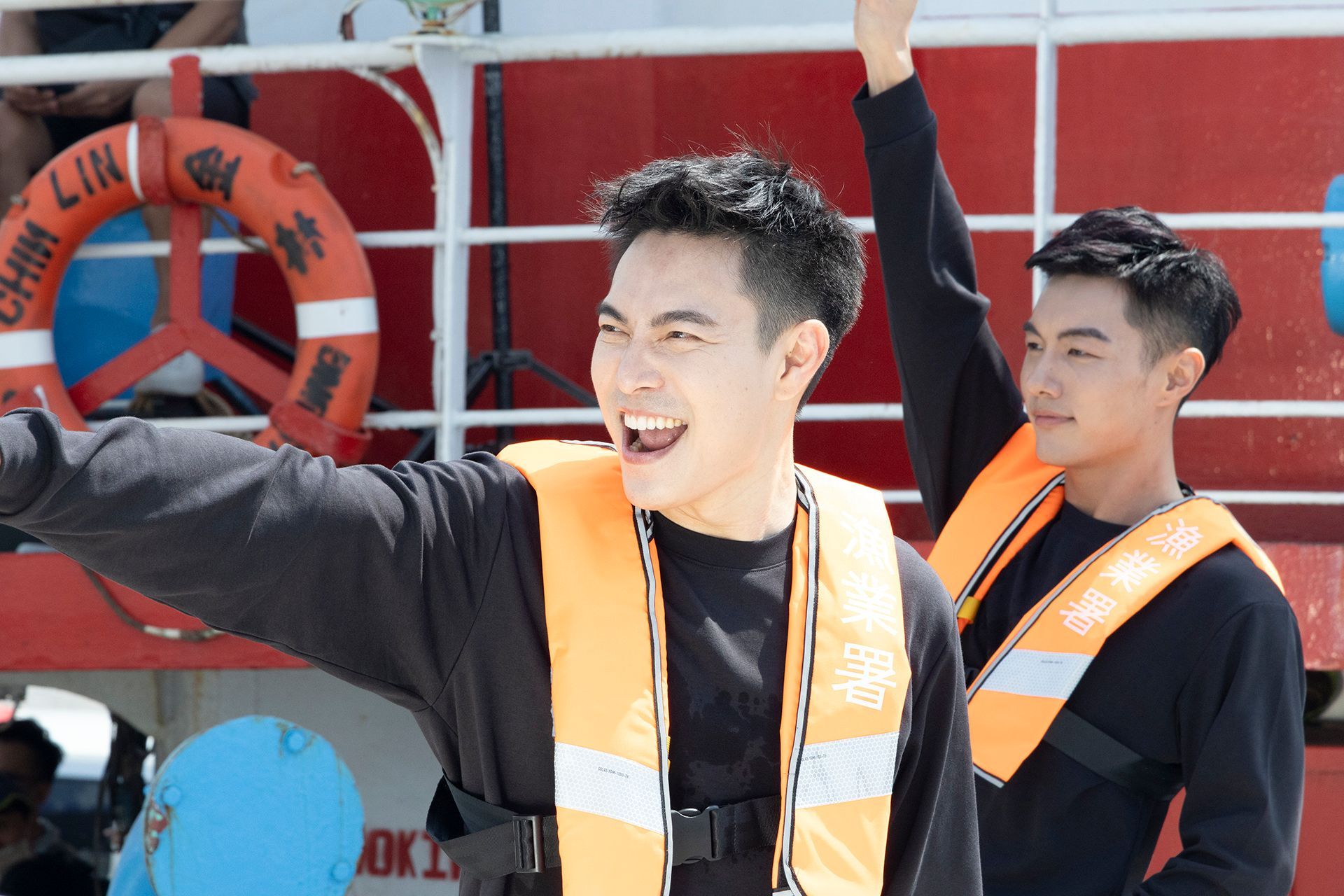 Two men on a boat wearing orange life vests, one smiling and pointing, the other raising a hand.