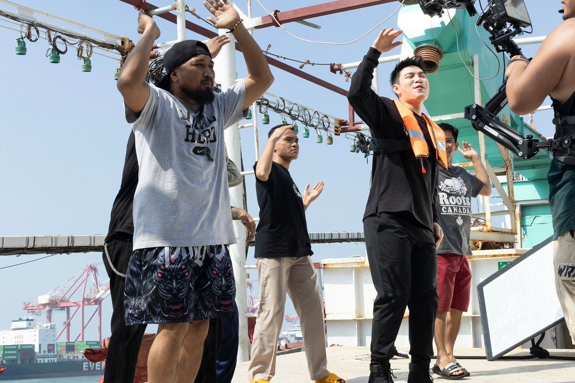 People on a boat with raised arms, filming. A camera operator is visible. Outdoors, bright day.