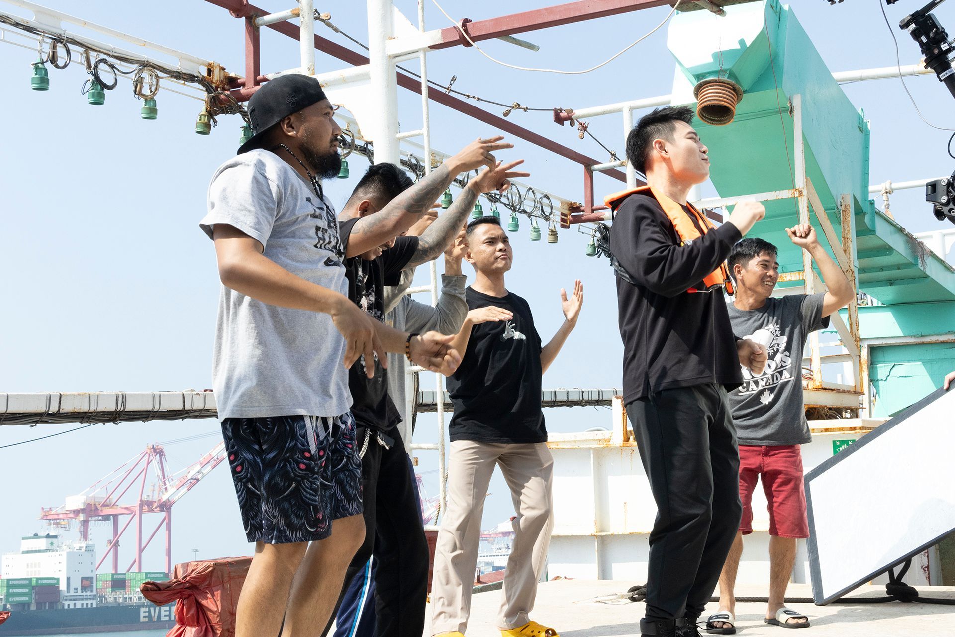 Five people dancing on a boat deck, hands raised. A harbor with cranes is in the background.