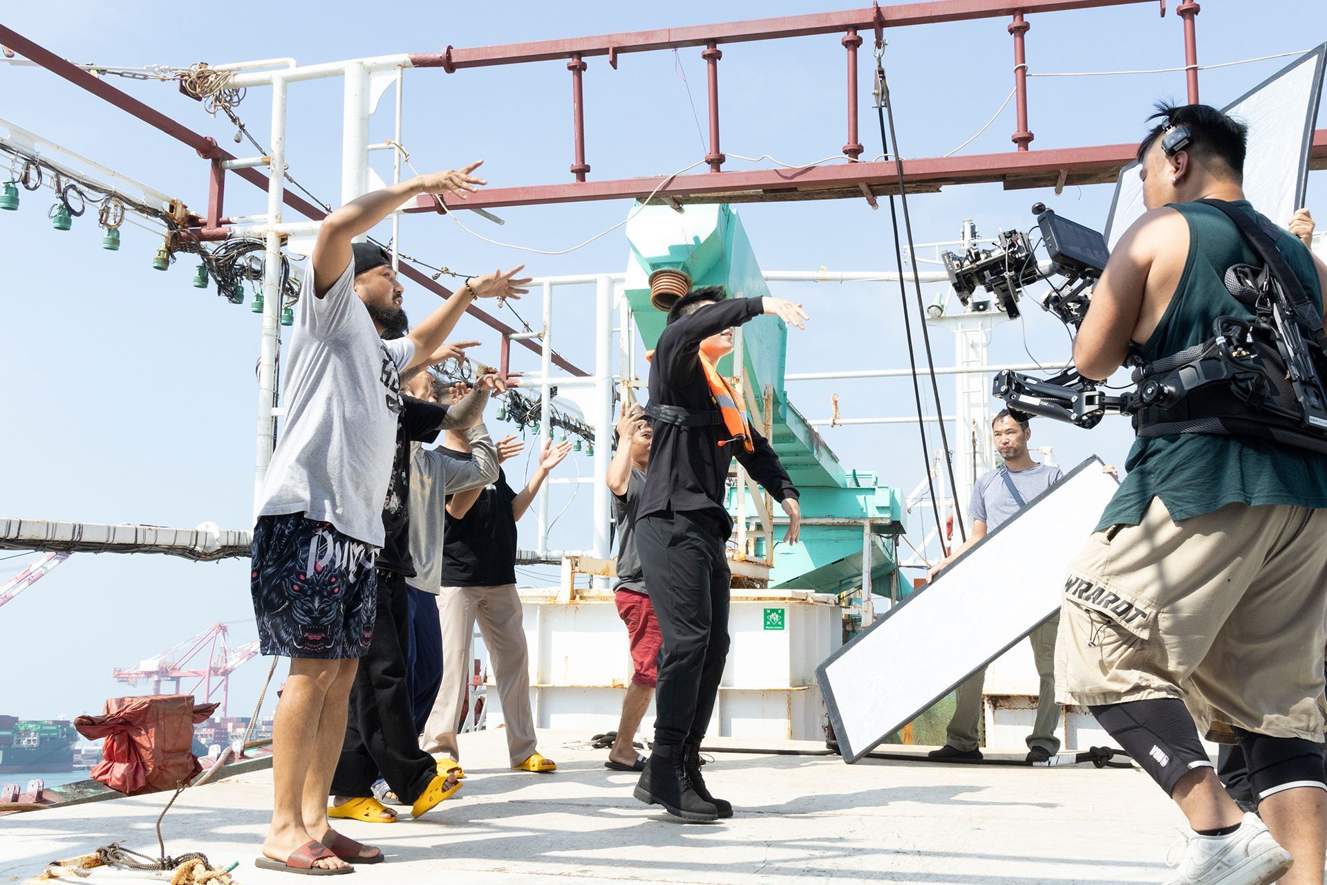Filming a scene on a boat deck; crew members with camera, lights, and actors in action, under blue sky.