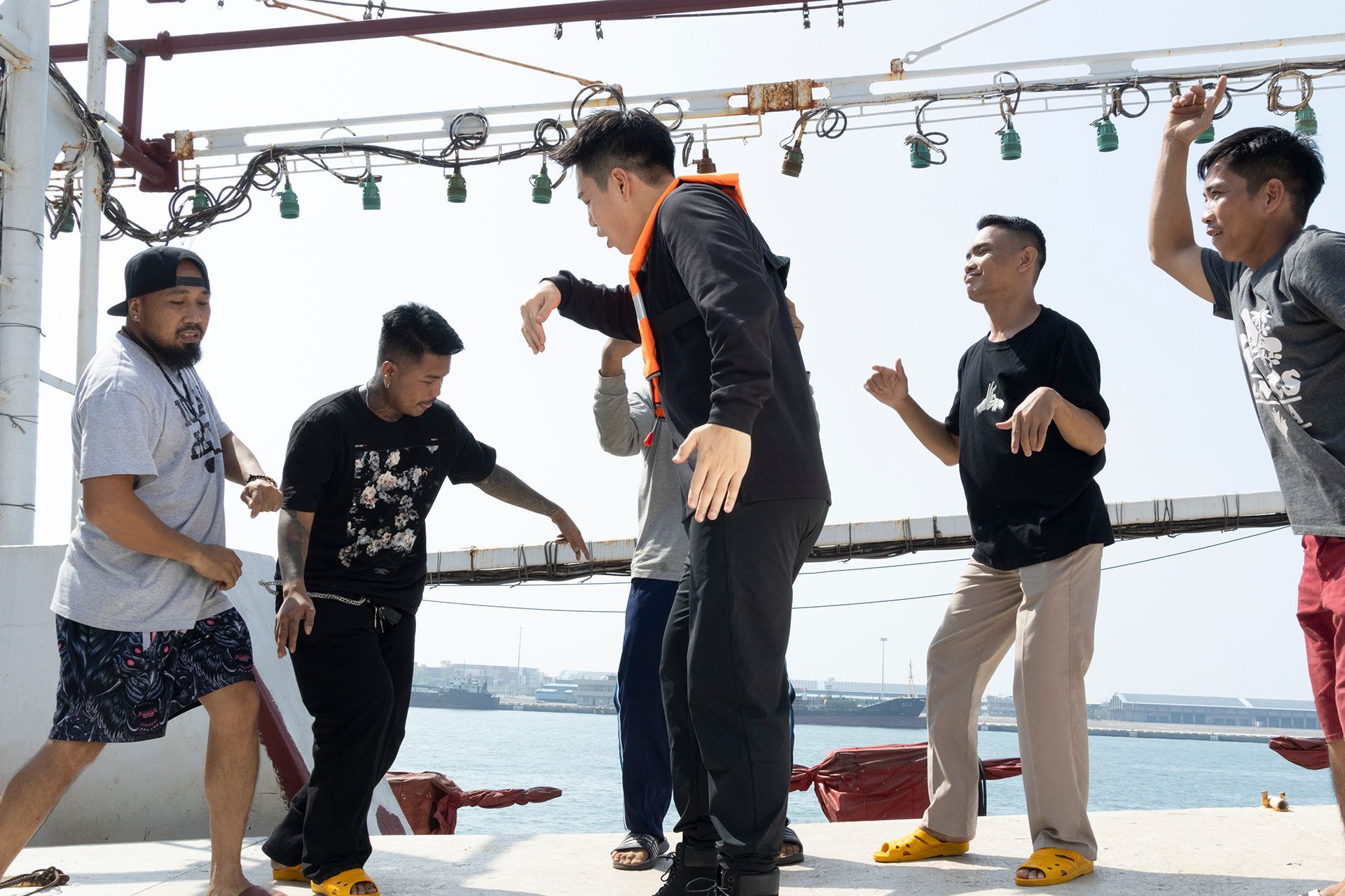 Men dancing on a boat deck in sunlight. Some wearing black, one in orange, gesturing with arms.