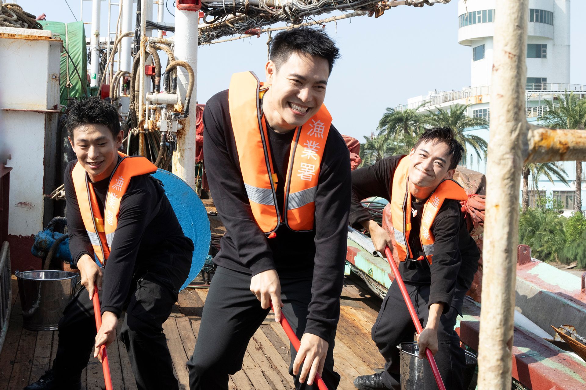 Three men on a boat, wearing life vests, smiling, and holding cleaning tools.