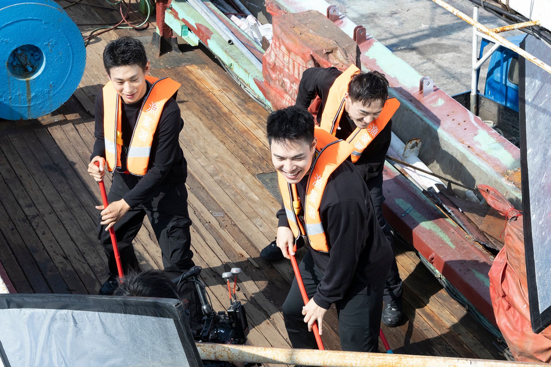 Four people wearing life vests sweep a boat deck; sunlight brightens the scene.