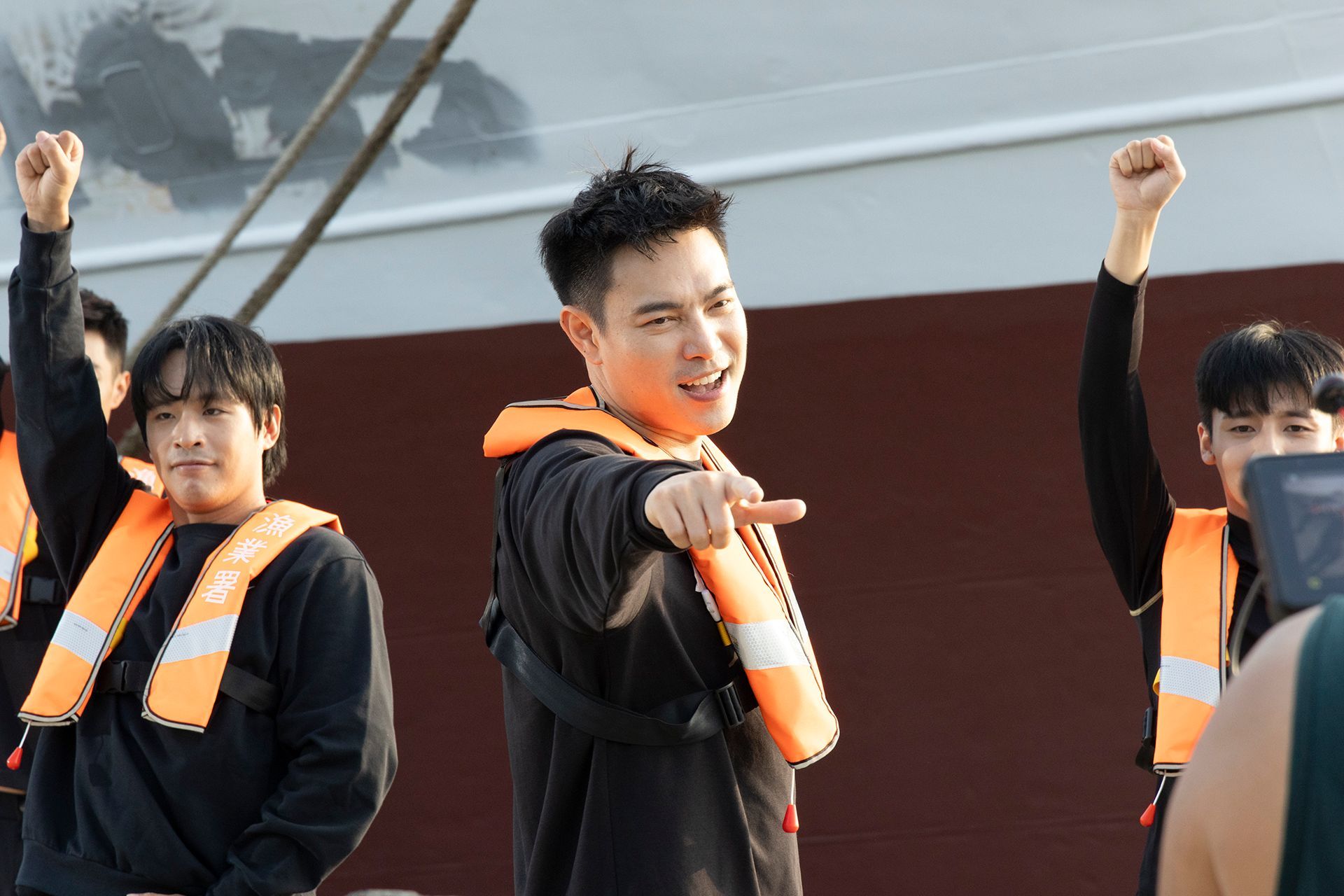 People in life vests point forward with fists raised near a large boat.