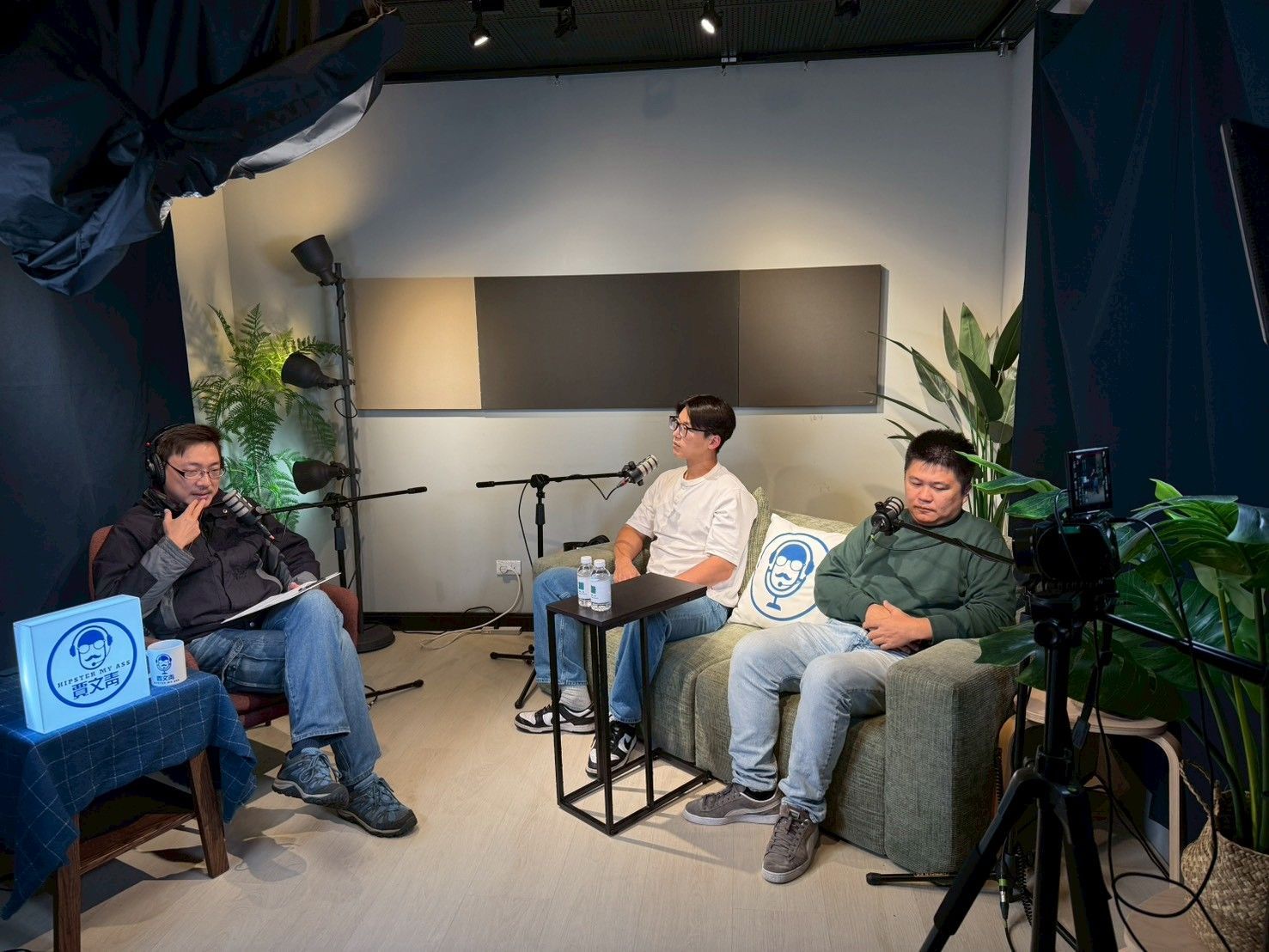 Three people in a podcast studio; two on a couch, one at a table. Microphones, plants, and a camera are visible.
