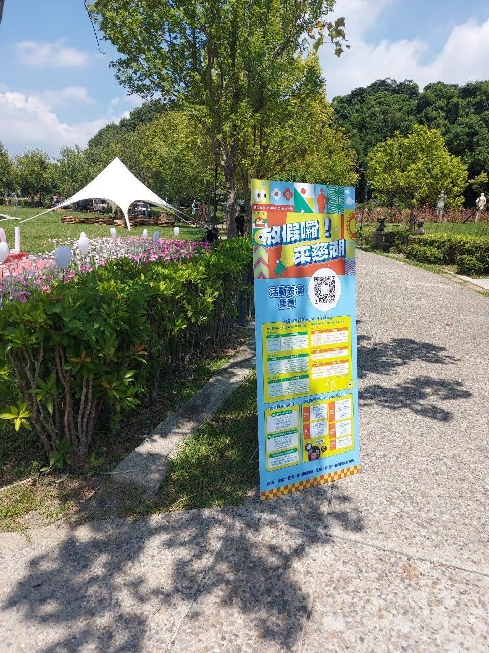 Signboard in a park with text and QR code; a white tent and greenery in the background.