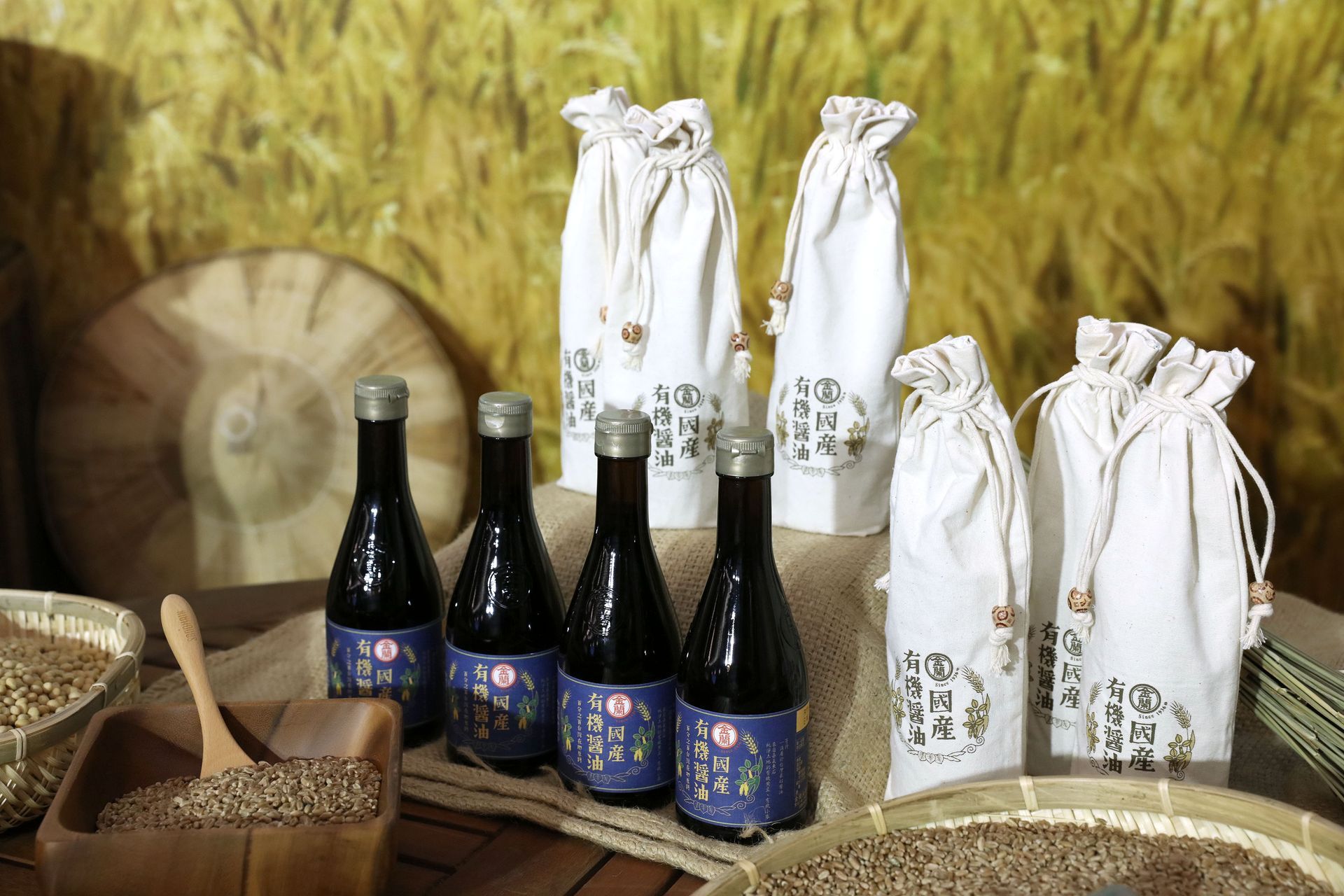 Bottles of dark liquid with blue labels and covered sacks on a table with grains and wheat background.