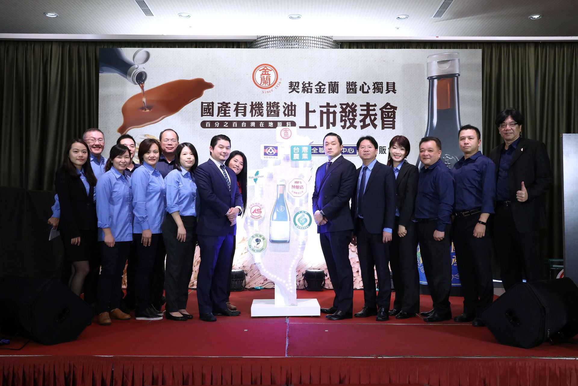 Group of people at a presentation event, standing on a stage with a logo backdrop.