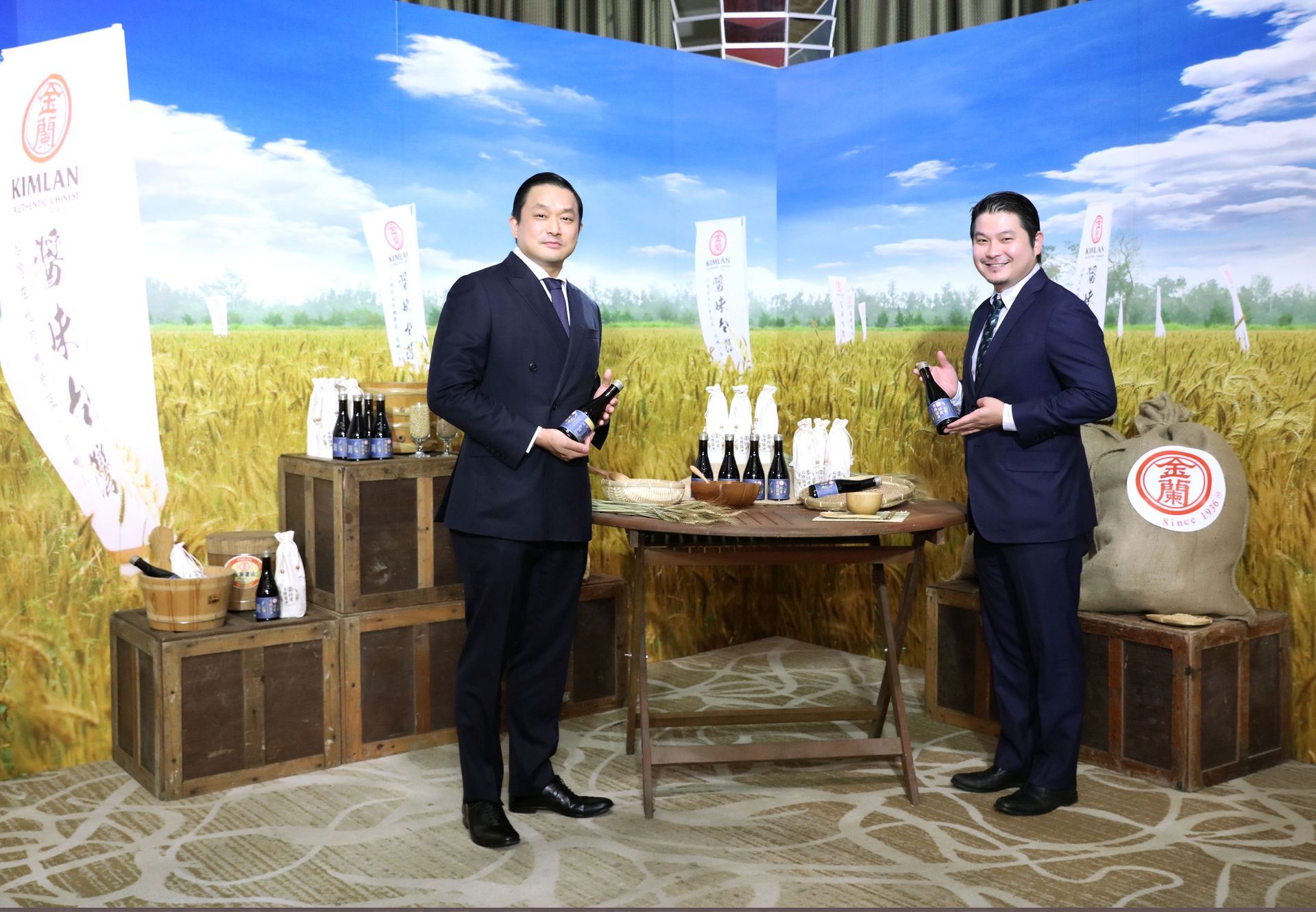 Two men in suits holding bottles stand at a table with drinks, in front of a harvest backdrop.