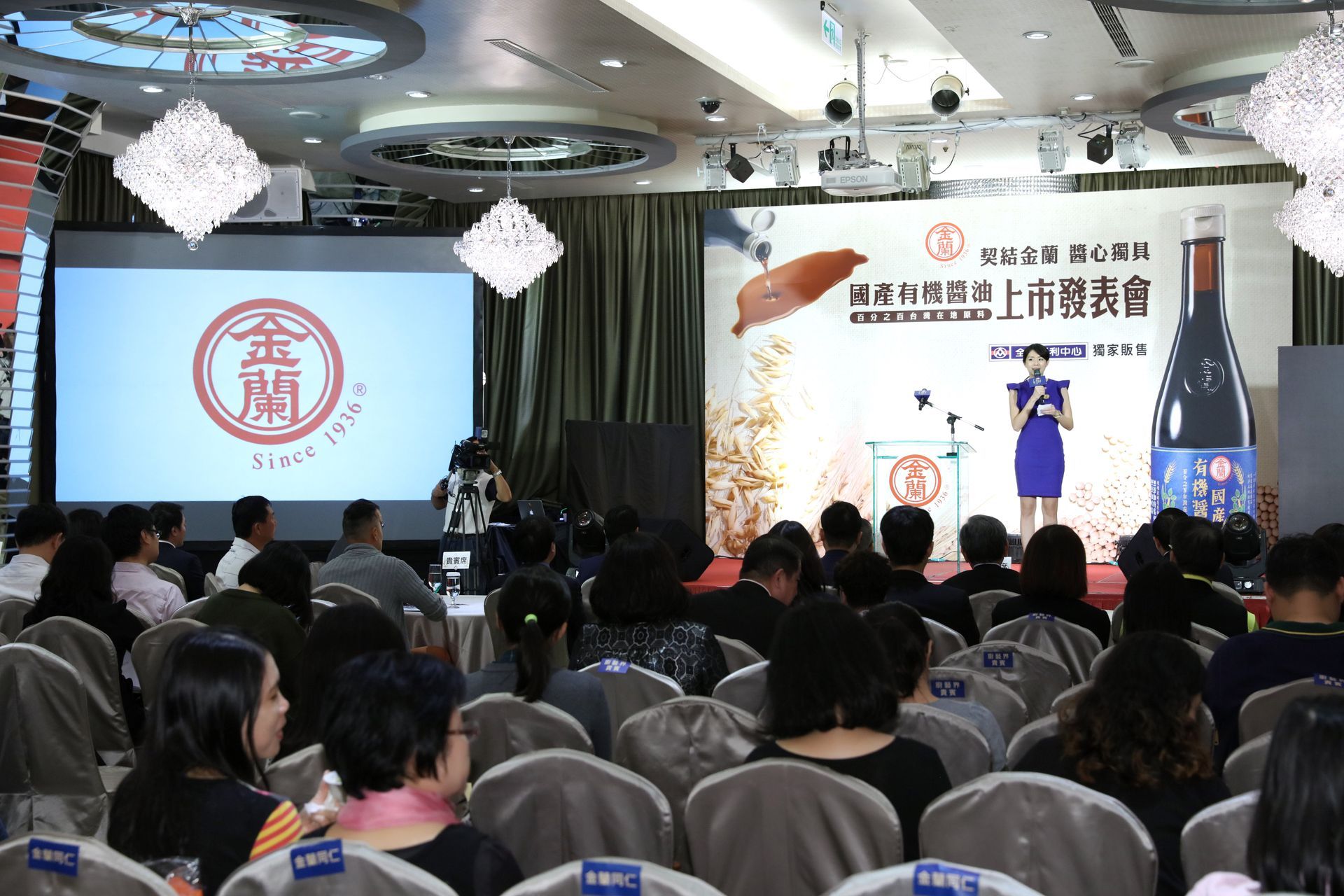 Event with a speaker at a podium, large screen with logo, audience seated in rows.