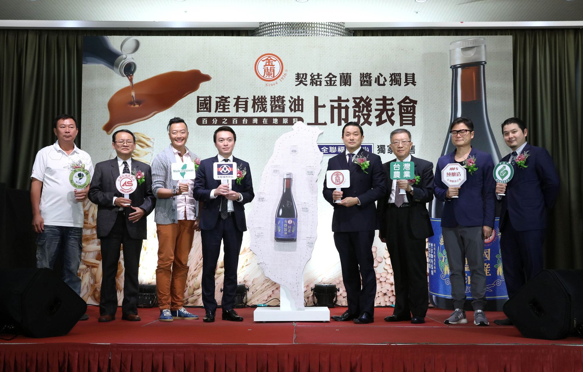 Group of men at a product launch event, standing in front of a backdrop, holding awards, red carpet.
