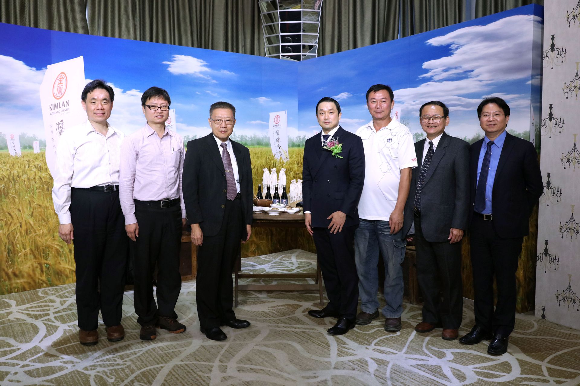 Group of eight men posing in front of a backdrop with a landscape scene.