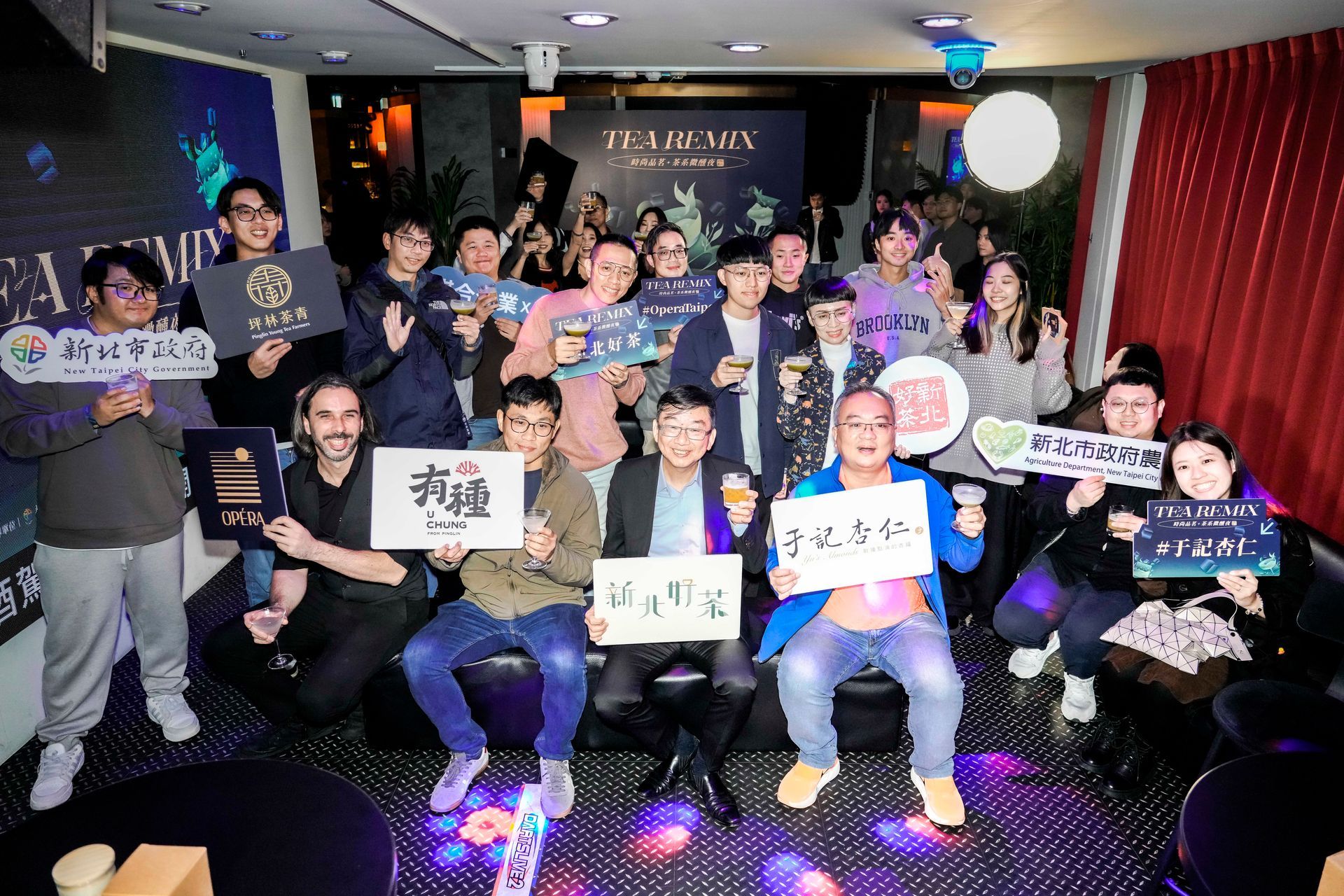 Group of people at an event, holding signs, some with drinks, smiling. Dark interior, possibly a bar.