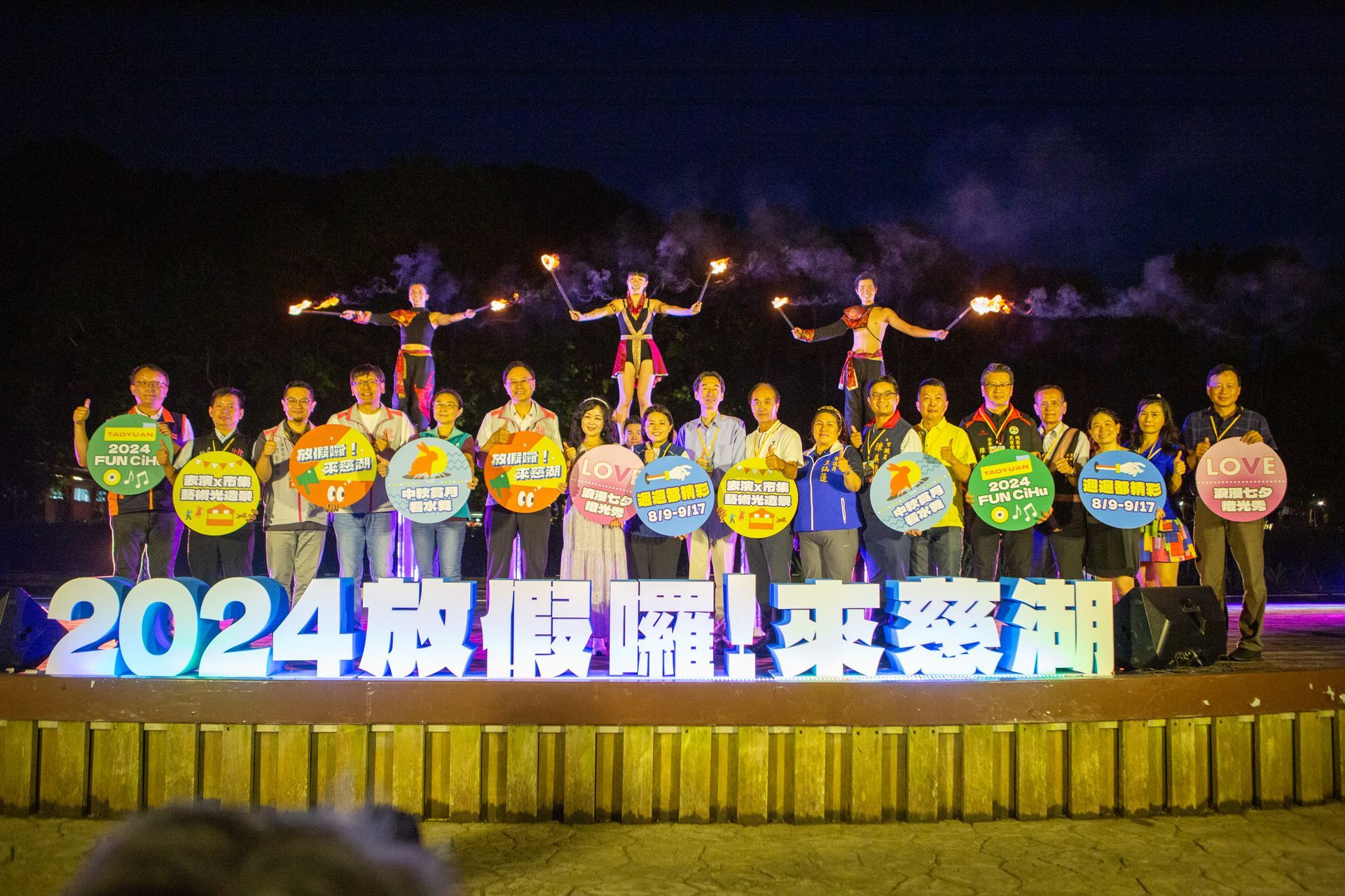 Group of people on stage with signs, fireworks performers overhead, nighttime outdoor event.