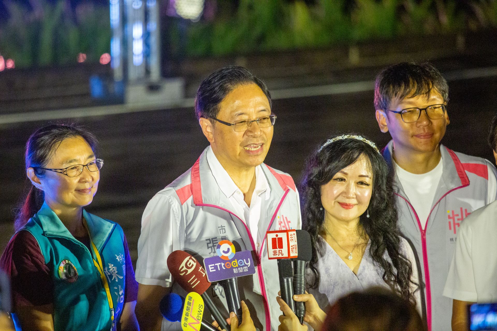 People with microphones in front, speaking outside at night.