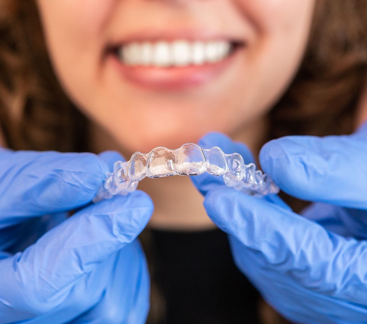 Gloved hands holding a clear dental aligner in front of a smiling mouth