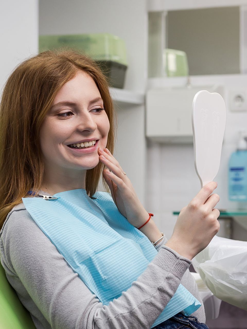 A woman is sitting in a dental chair looking at her teeth in a mirror.