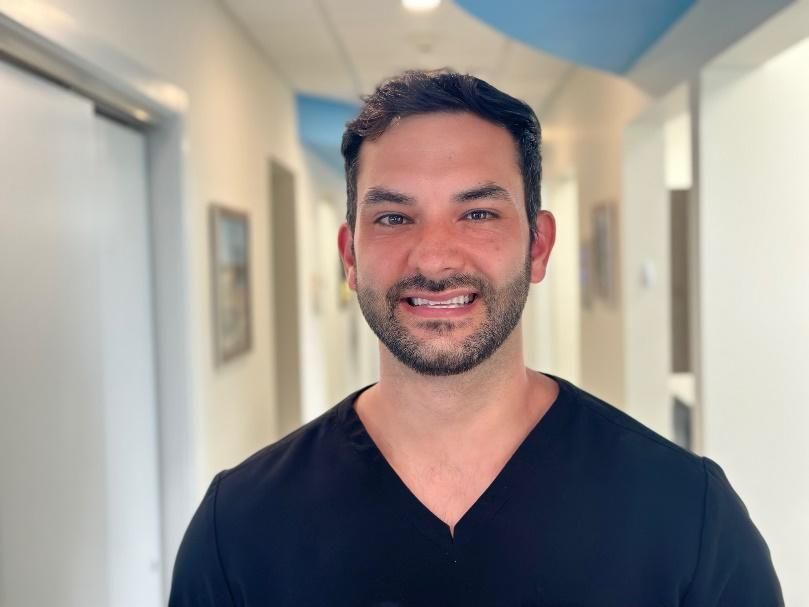 A man with a beard is wearing a black scrub top and smiling for the camera.