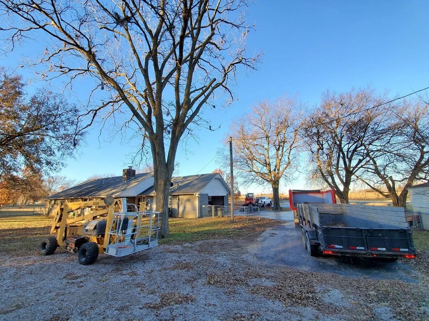 Backyard with a house, leafless trees, a trailer, and a utility vehicle on a gravel drive