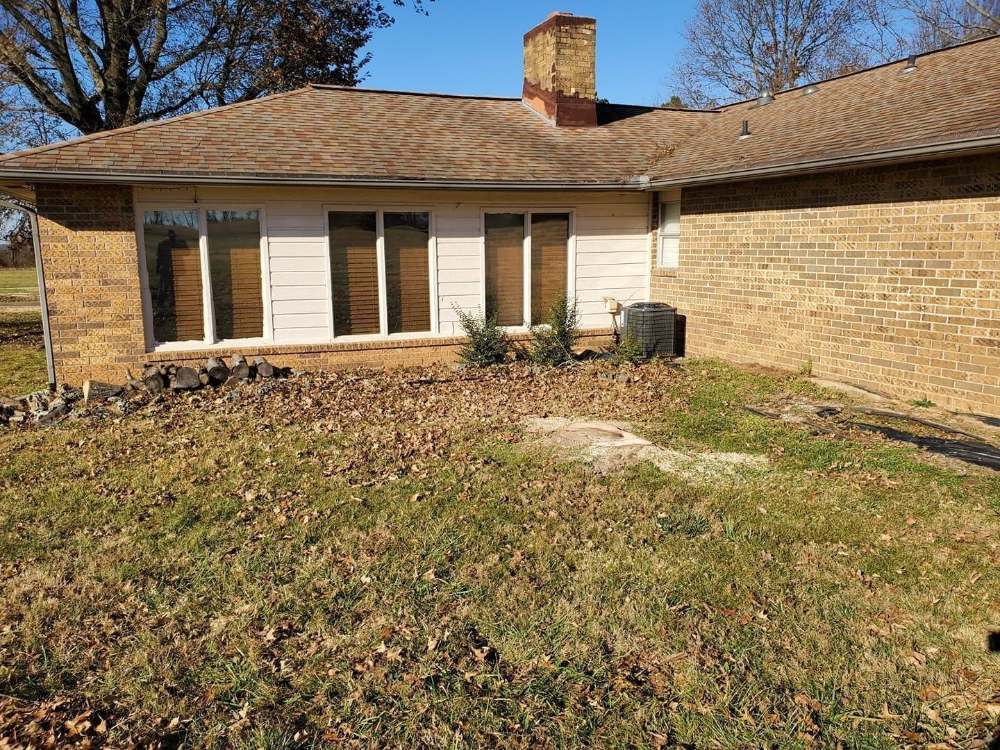 Single-story brick house with tan roof, front windows, and a grassy yard in sunlight