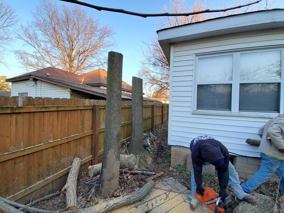 Two workers digging near a house and wooden fence beside exposed posts and debris