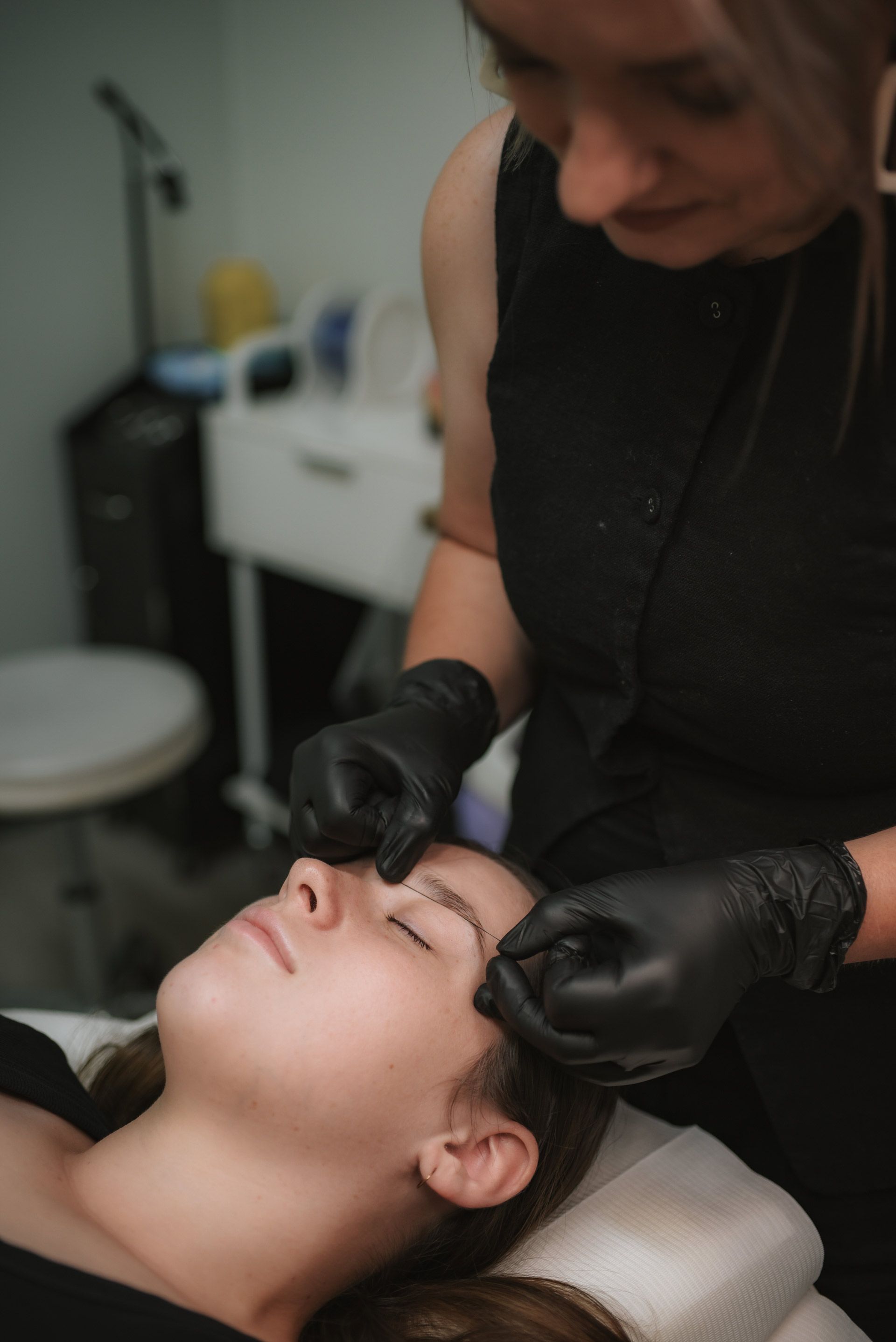 Woman getting eyebrow procedure; technician wearing black gloves and outfit working close up on the client's face.
