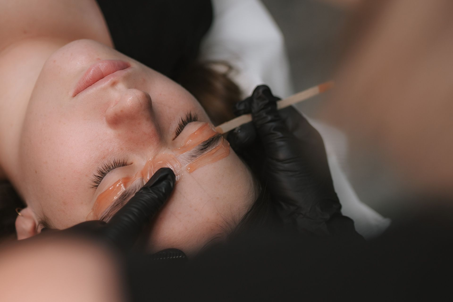 Woman receiving eyebrow waxing at a salon, gloved hands applying wax.
