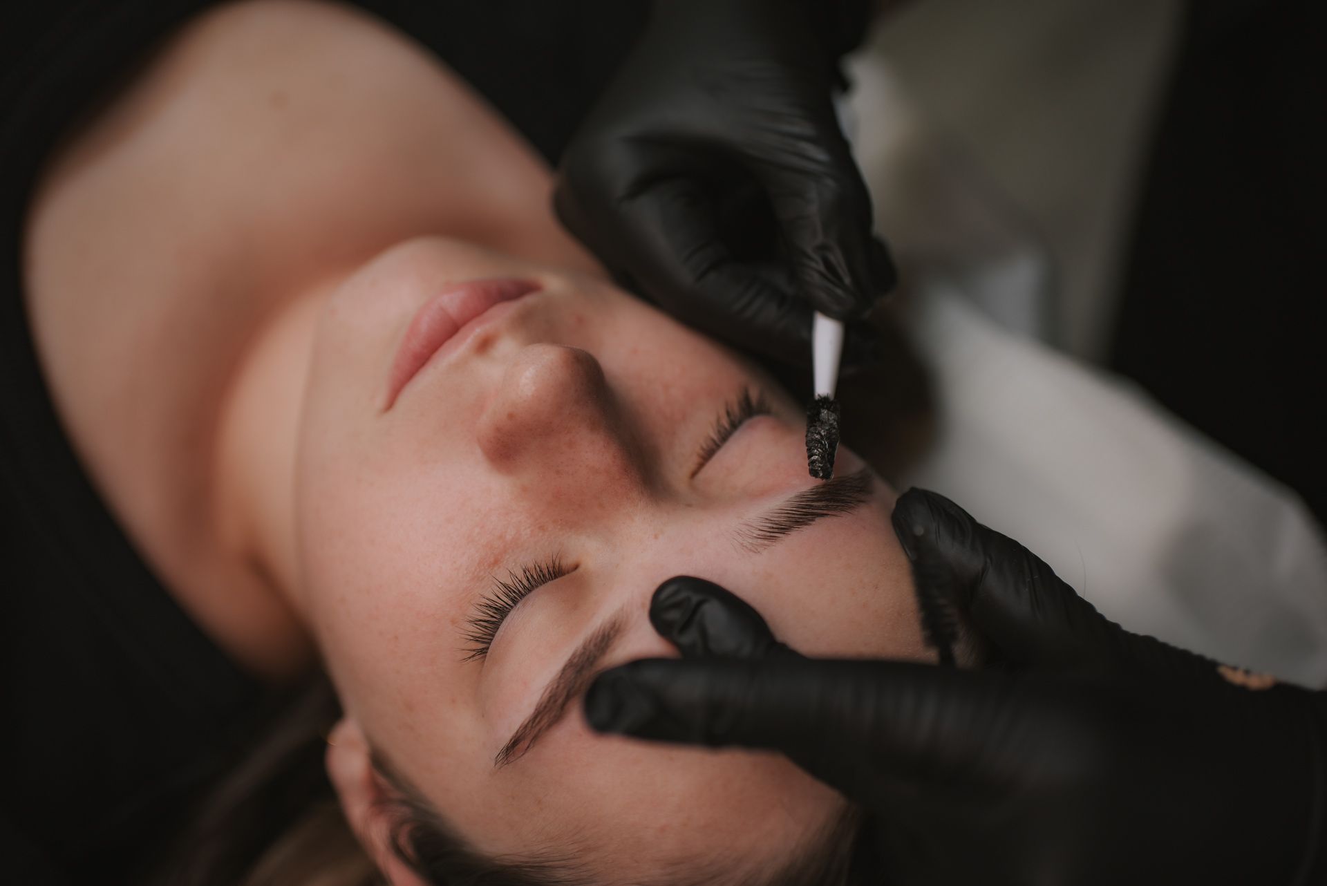 Person receiving an eyebrow tattoo, hands with black gloves holding a tool, white setting.