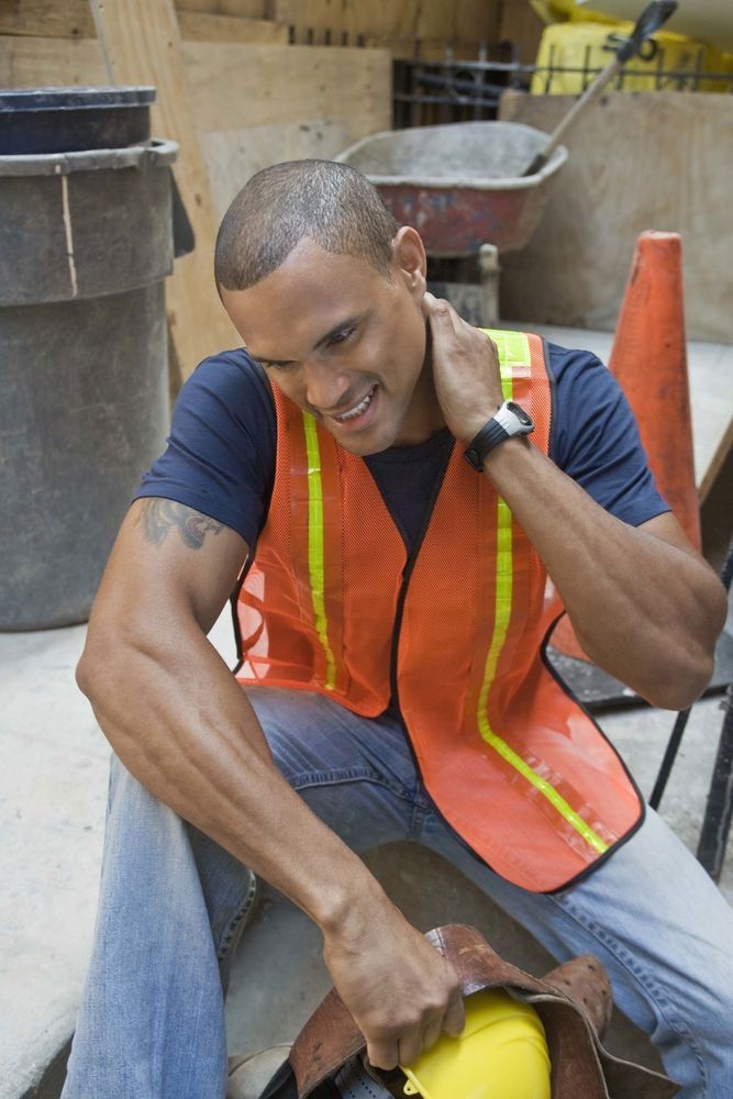 Construction worker with orange vest, rubbing neck, seated in a construction zone.
