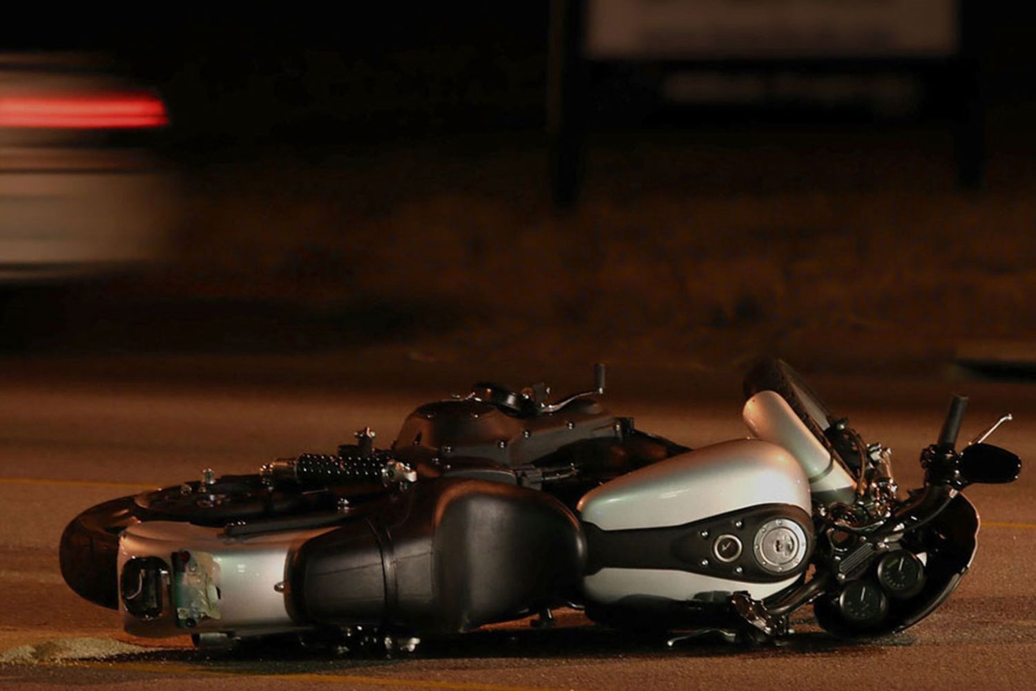 Motorcycle lying on its side on a road at night, with blurred car lights passing in the background.