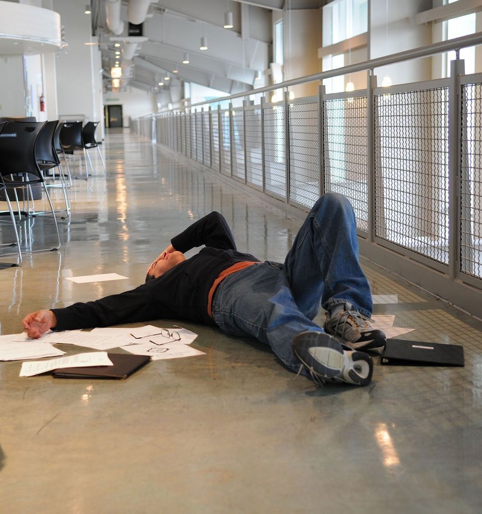 Man lying exhausted on floor of a modern hallway, papers scattered, laptop nearby.