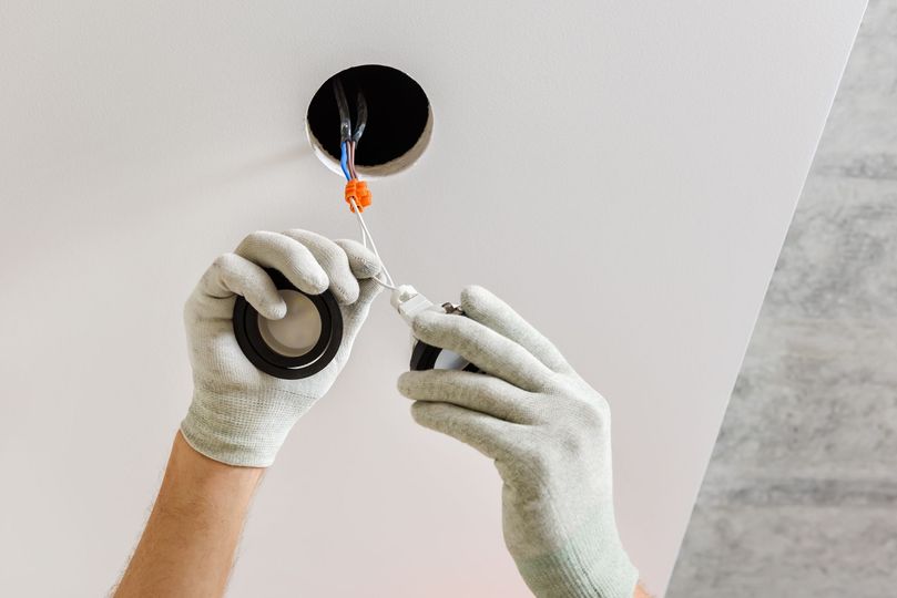 Hands in work gloves installing an electrical fixture in a white ceiling.