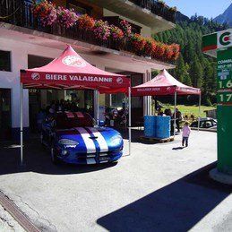 Une voiture de sport bleue est garée sous une tente rouge devant un immeuble.